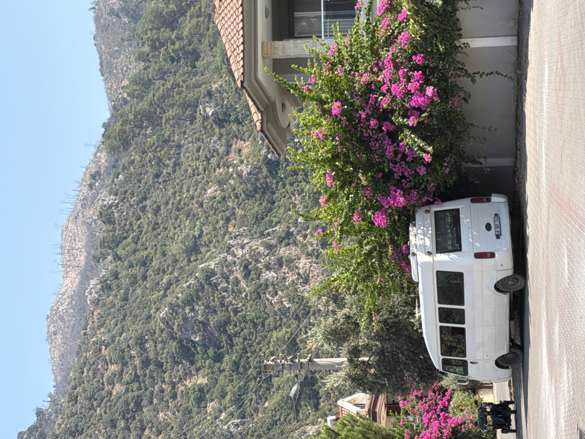 In Marmaris, Turkey, a white minibus is parked on a paved road in front of a wall covered in vibrant pink bougainvillea. The minibus has its rear facing the camera, and a license plate that reads "48 BAY 742." To the left of the minibus, a black electric scooter or small cart is parked. Behind the wall and bougainvillea, a house with a tiled roof and balconies is visible. In the background, a steep, green, and rocky mountainside rises under a clear blue sky. The overall scene depicts a sunny day in a coastal town.