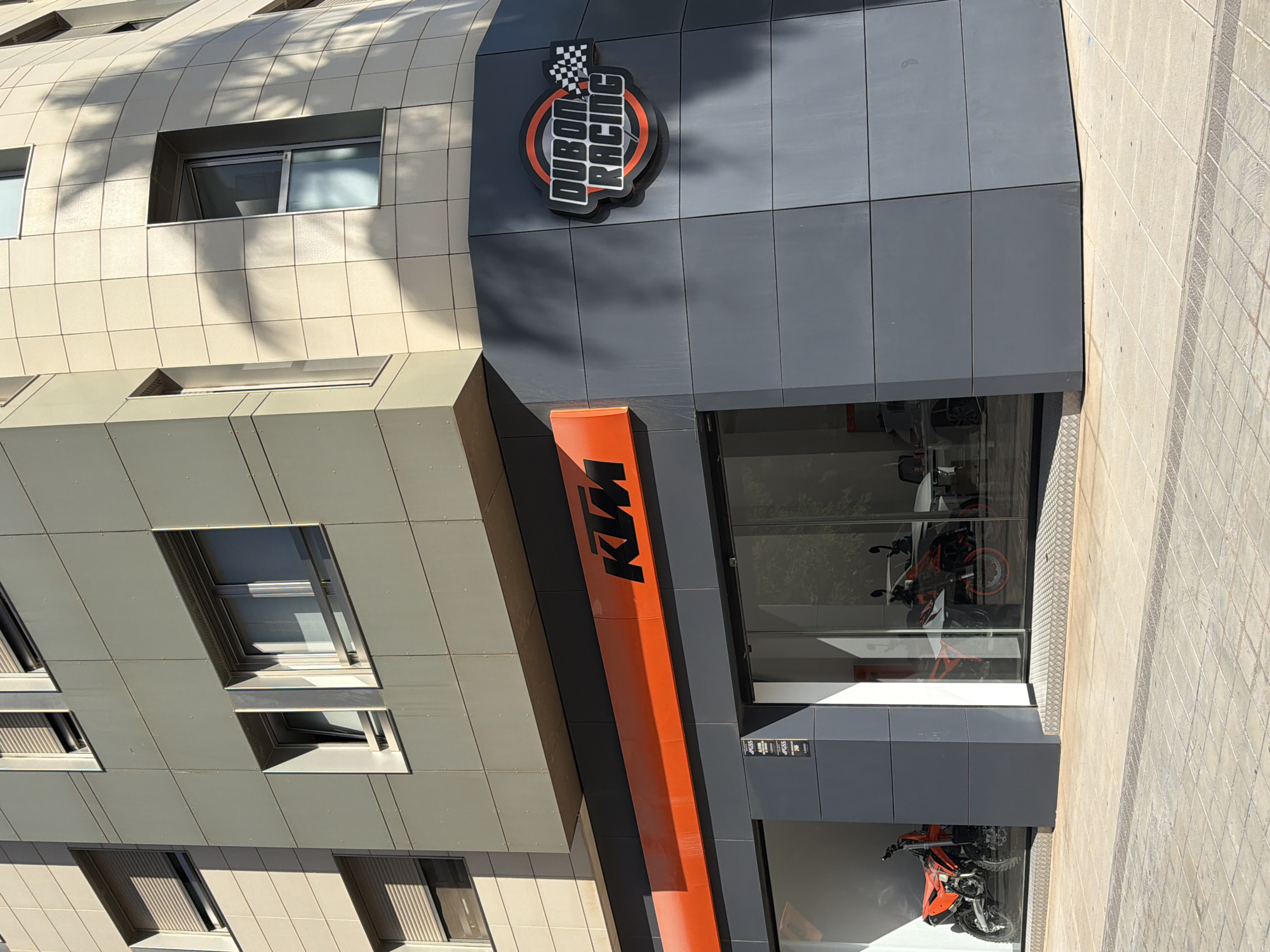 The image shows the exterior of a KTM dealership, "Dubon Racing," located in an urban environment. The building features a modern facade with grey and cream-colored tiled panels and large windows. Prominently displayed on the building are the "KTM" logo in orange and black, and a circular "Dubon Racing" logo with a checkered flag detail. Through the large glass storefront, several motorcycles, including what appears to be a red dirt bike and other KTM models, are visible inside the showroom. The pavement in front of the building is paved with light-colored tiles. Sunlight casts shadows from palm trees onto the building's facade.