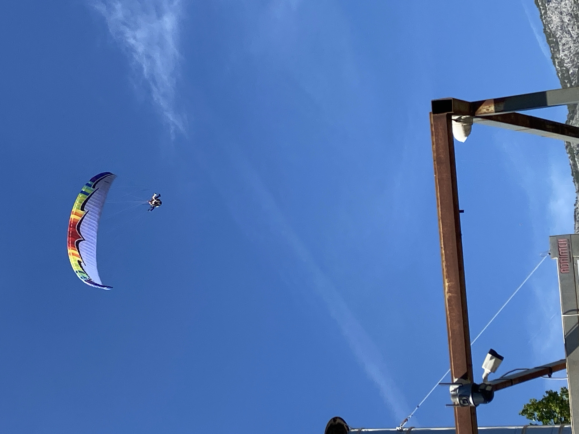 The image shows a paraglider in a clear blue sky. The paraglider is a colorful, curved wing with a person suspended below. The background is a clear, deep blue sky with a faint contrail. In the foreground, parts of a metal structure are visible, including what appear to be security cameras. A sign that reads "ADRIATIC II" is visible on the right side of the image. The location is Boreti, Montenegro.