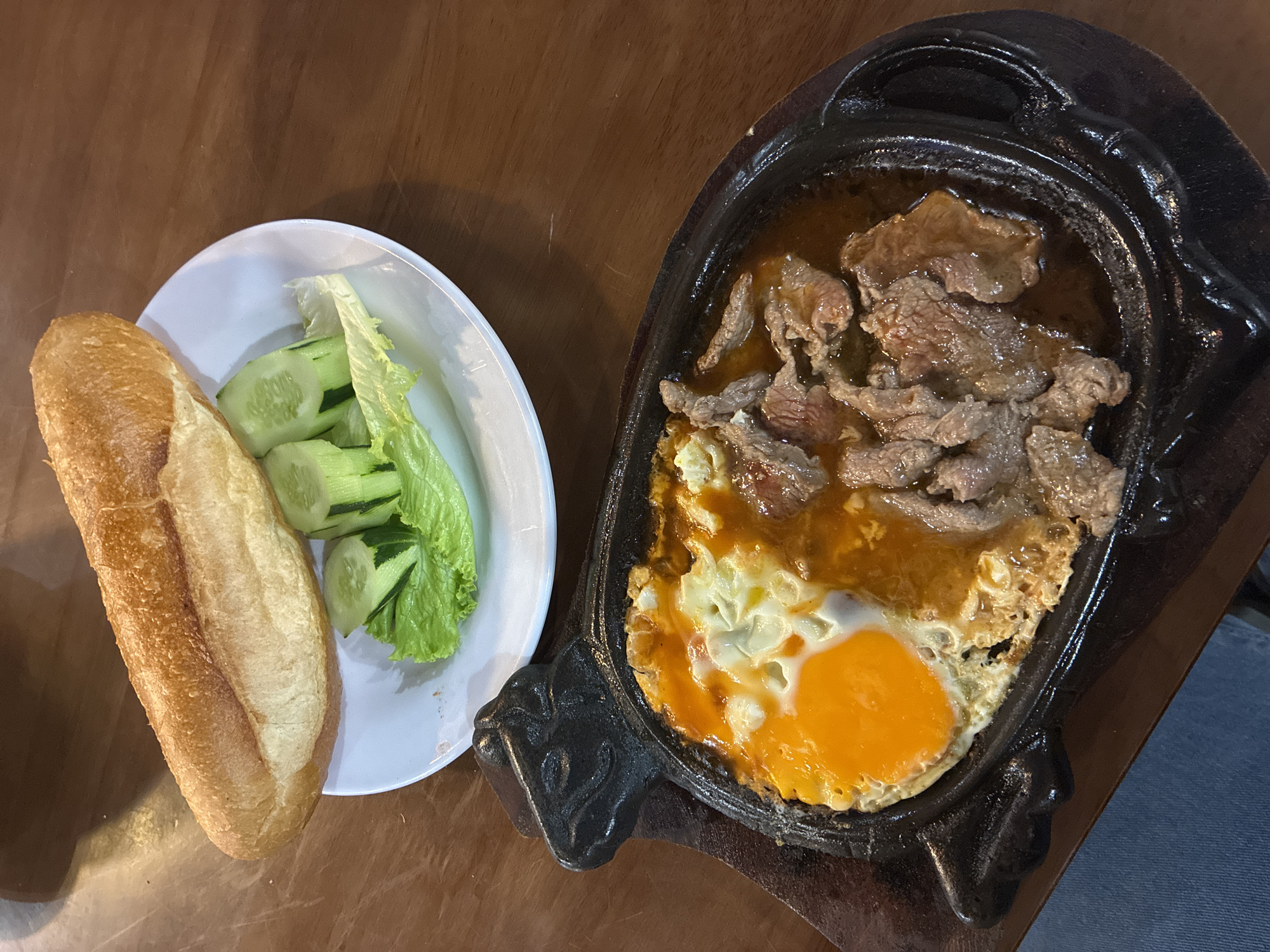 The image shows a wooden table with two dishes. On the left is a white plate with a baguette, sliced cucumbers, and lettuce. On the right is a cast iron skillet containing a fried egg and sliced beef in a sauce. This is likely a serving of Bò Né, a popular Vietnamese street food dish often found in Đà Nẵng. The scene is likely in a restaurant or food stall in Đà Nẵng, Vietnam.