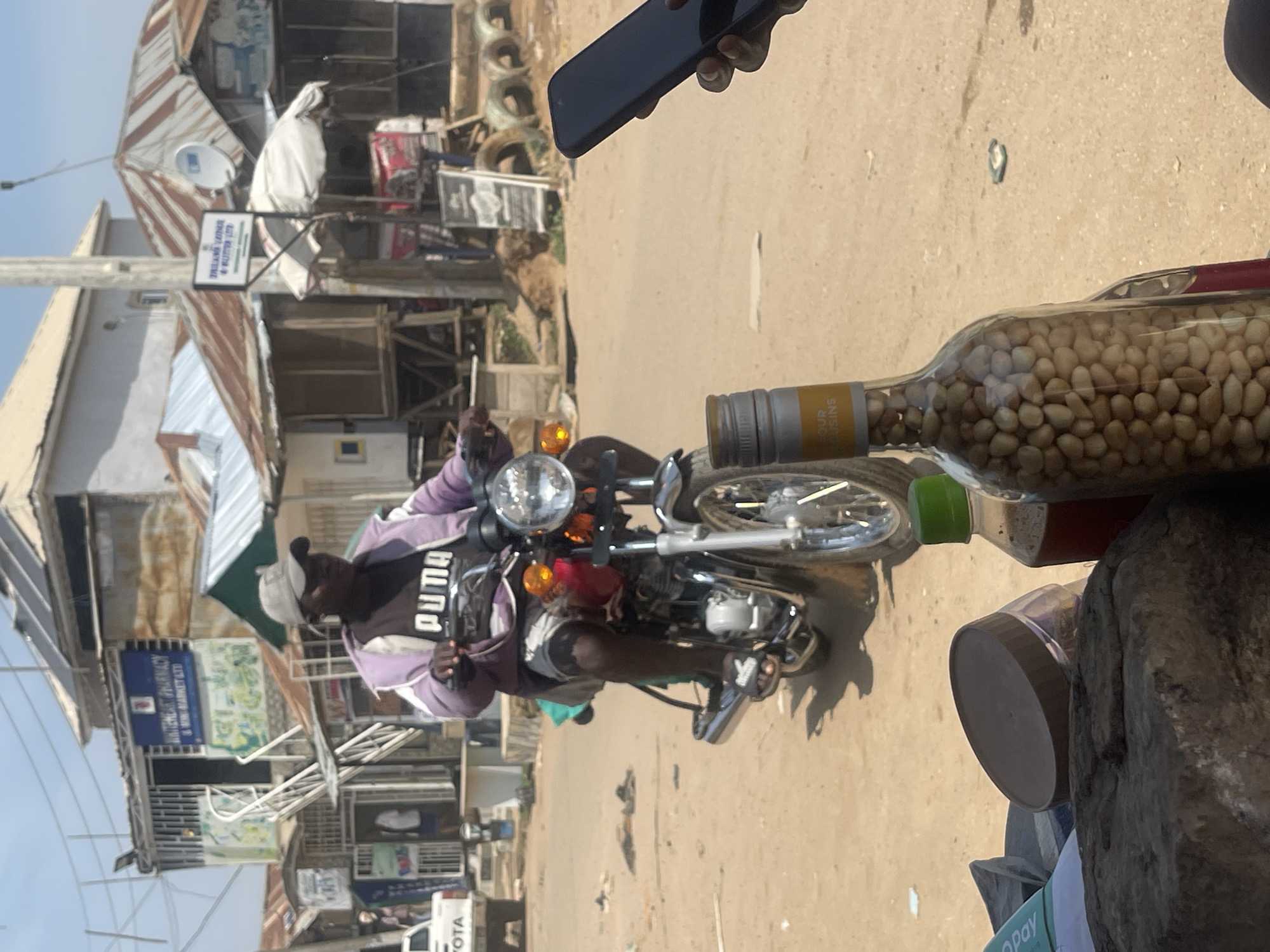 This is a medium shot of a man riding a motorcycle in what appears to be a marketplace in Abuja, Nigeria. The man is wearing a purple jacket over a black shirt with "PUMA" written on it, and a grey baseball cap. He is looking forward, hands on the handlebars. The motorcycle is red and appears to be a common model for this region.
The background shows a street scene with various small shops and stalls. There are buildings with corrugated iron roofs, typical of urban areas in Nigeria. Signs are visible, including one that reads "WHITEHEART PHARMACY & MINI MARKET LTD". To the right, a person's hand is holding a dark-colored smartphone, partially obscuring the view of the street. In the foreground, out of focus, are a glass bottle filled with peanuts and another bottle containing a dark liquid. The ground is sandy and uneven, suggesting a dirt road or unpaved area. The sky is clear and bright, indicating it is daytime. The overall atmosphere is that of a busy, functional marketplace.