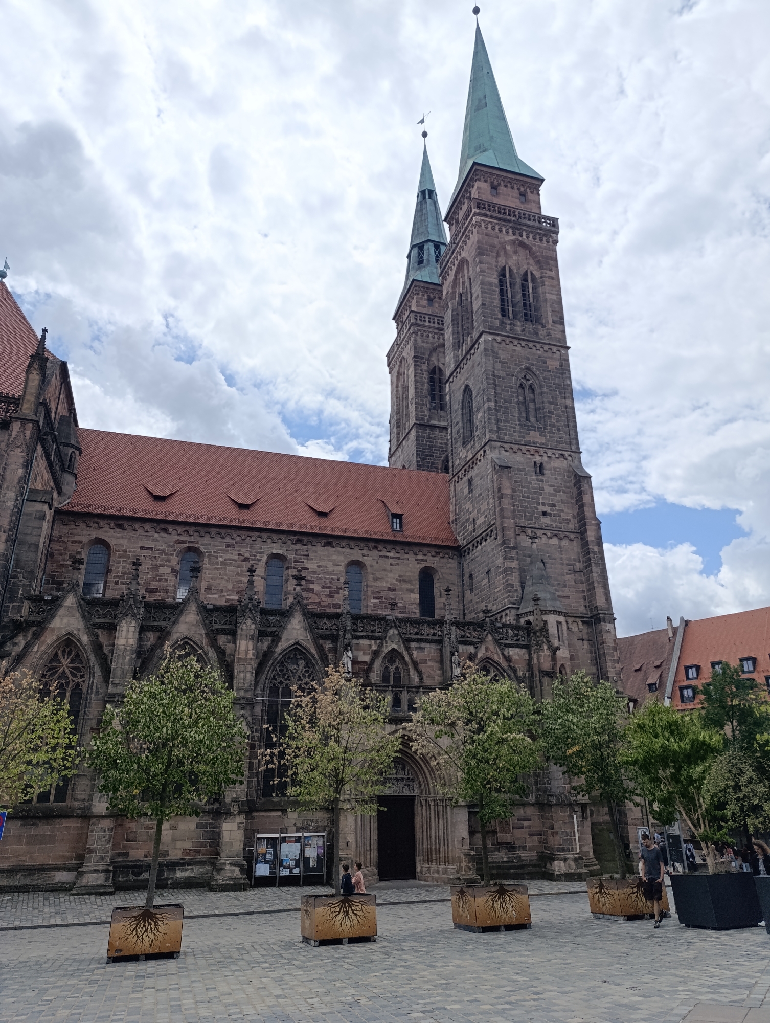 The image depicts Saint Lorenz Church, a prominent Gothic church located in the city of Nuremberg, Germany. The church's facade is constructed from dark stone, with a large red tiled roof. It features two tall spires, one significantly larger than the other, both topped with green copper roofs. Intricate architectural details, including pointed arch windows and ornate stonework, are visible on the church. In front of the church, the ground is paved with cobblestones. Several trees are planted in square planters adorned with a root design. Two individuals are standing near the entrance of the church, and a third person is walking across the cobblestone plaza. The sky is filled with scattered clouds, indicating an overcast day.