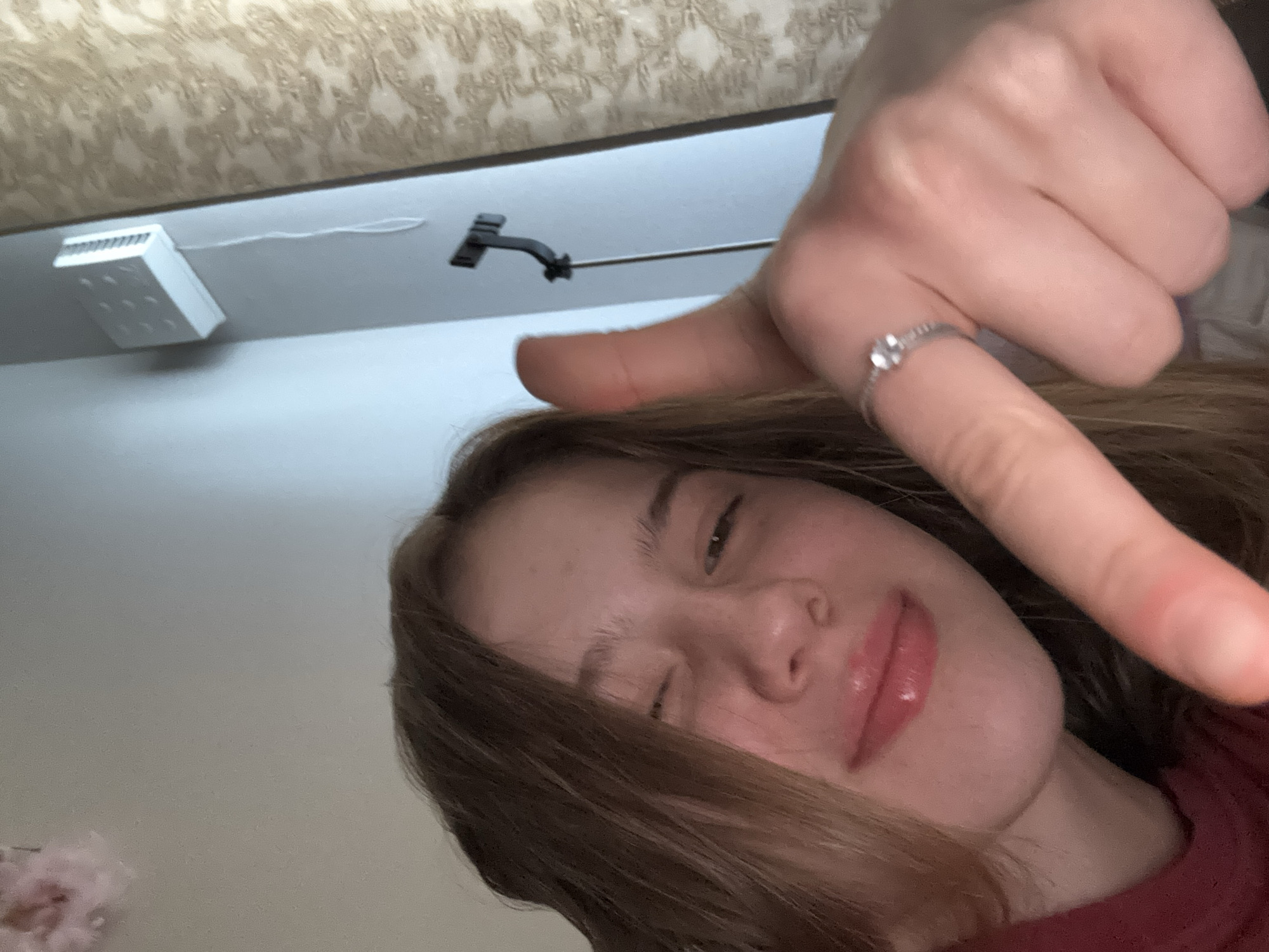 A young woman with light brown hair is taking a selfie in a room. She is winking and giving a thumbs-up gesture towards the camera, while also pointing downwards with her index finger. She is wearing a red shirt and a silver ring with a small stone on her left index finger. Her lips are glossy. In the background, a white vent is mounted on a light blue wall, and a patterned curtain is visible on the right. A selfie stick is attached to the wall behind her. This scene is taking place in Vsevolozhsk, Russia.