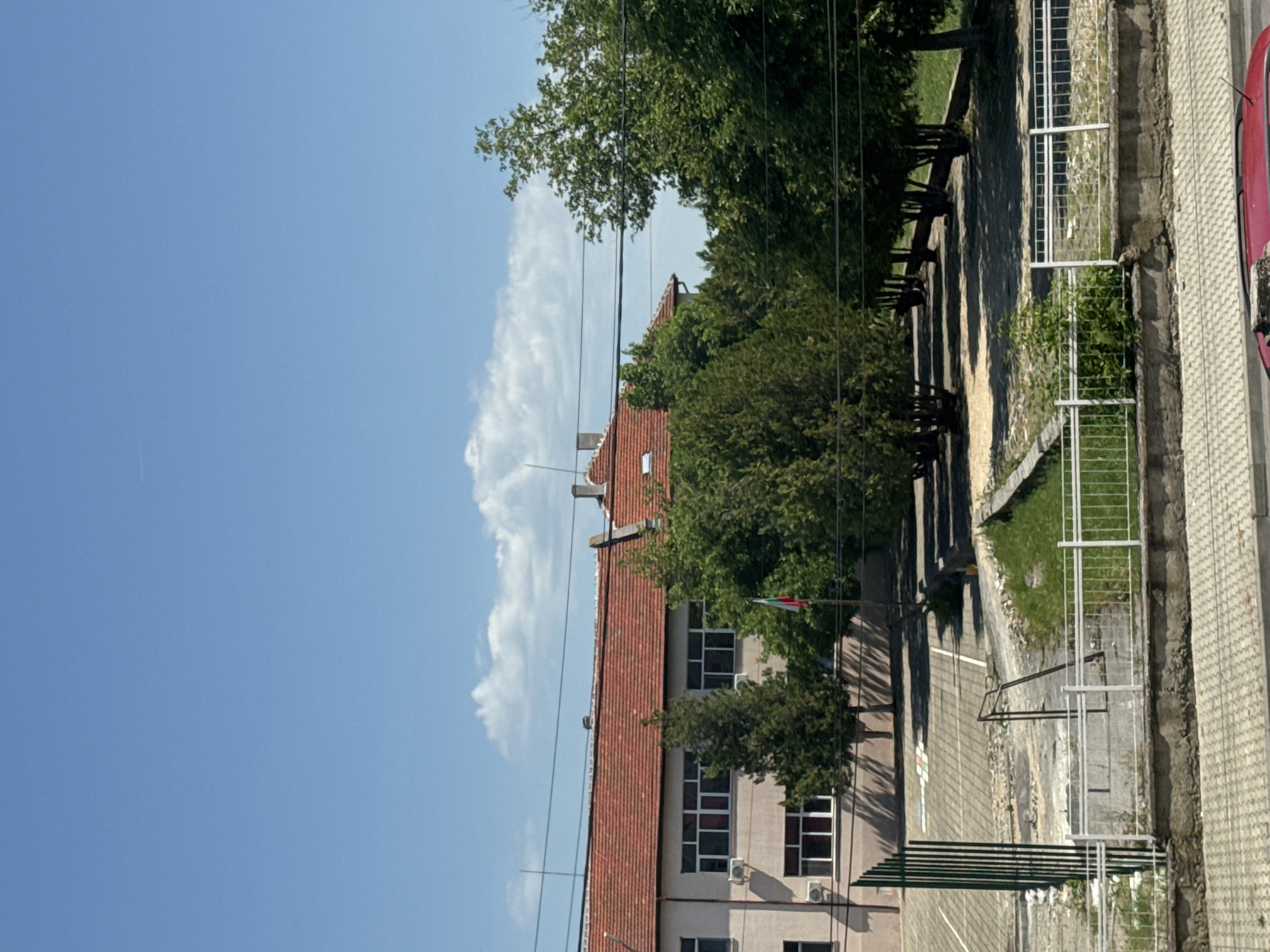 This image shows a building in Unknown, Unknown. The building has a red tiled roof and is surrounded by trees. A Bulgarian flag is visible, flying from a pole near the building. There is a fence and a sidewalk in front of the building. The sky is blue with white clouds.