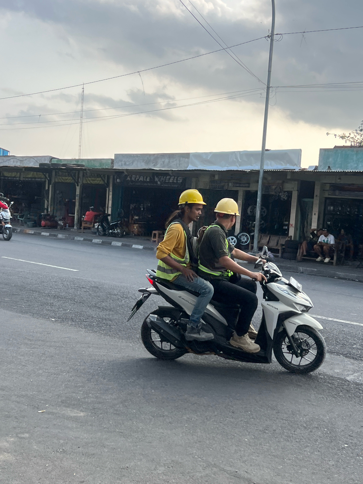 Two men wearing yellow hard hats ride a white Honda scooter on a paved road in front of a row of storefronts. The man in the front is wearing a dark green t-shirt and black pants. The man behind him is wearing a yellow long-sleeved shirt and blue jeans. Both are wearing high-visibility vests over their clothing. The storefronts appear to be shops, some with open fronts displaying goods like tires and wheels. A man in an orange shirt is seated inside one of the shops. Several other people are seated outside another shop in the background. The sky is overcast with some clouds. The scene is likely a commercial area on a street in Yogyakarta, Indonesia, possibly a business district or a market area known for auto parts.