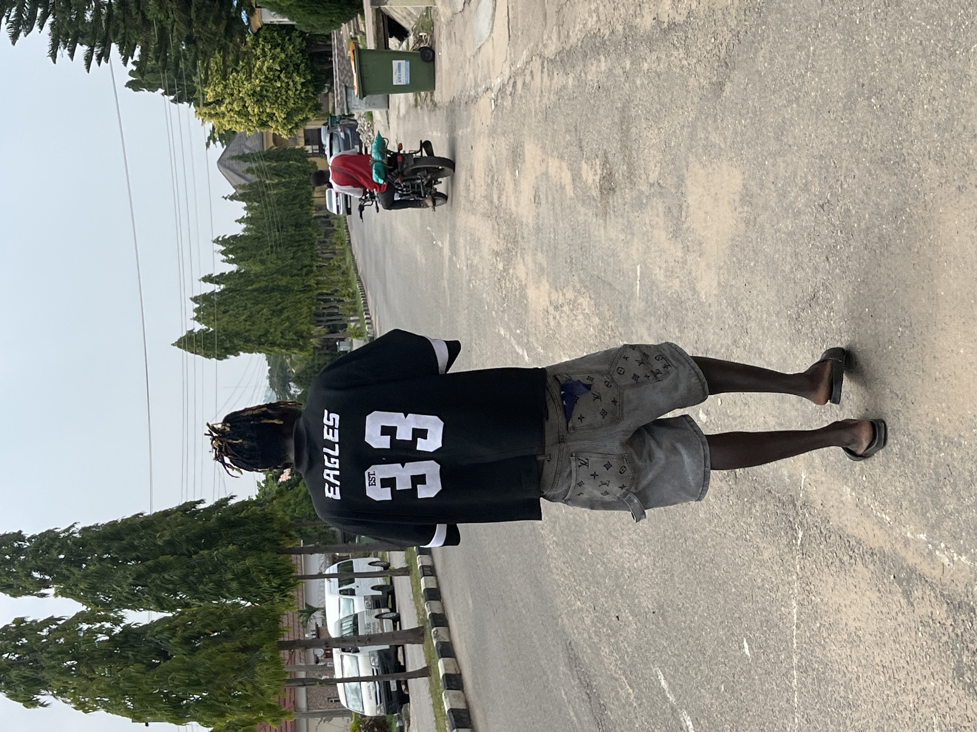 In a city street in Abuja, Nigeria, a young Black man with dreadlocks walks away from the camera. He is wearing a black t-shirt with white lettering that says "EAGLES EST. 33" and grey denim shorts with a repeating pattern. He is wearing black slides. The road is paved and has a white line marking the center. To the left of the road are several tall, slender trees and parked white vans. To the right of the road are more trees, a residential building with a dark roof, and a green trash bin. In the distance, a person in a red shirt rides a motorcycle away from the camera, carrying blue bags on the back. The sky is overcast.