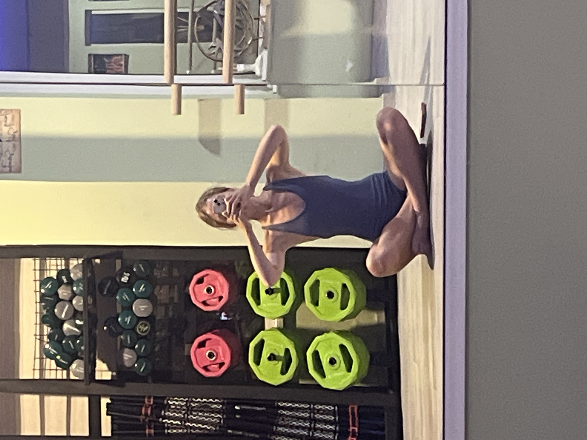 A woman is sitting cross-legged on the floor in front of a reflective surface, taking a mirror selfie with a mobile phone. She is wearing a dark blue sleeveless leotard. In the background, to her left, is a rack of gym equipment, including weight plates in pink and lime green, and what appear to be dumbbells or kettlebells on higher shelves. To her right, visible through a glass door, is a ballet barre and other gym equipment. The environment appears to be a gym or fitness studio.