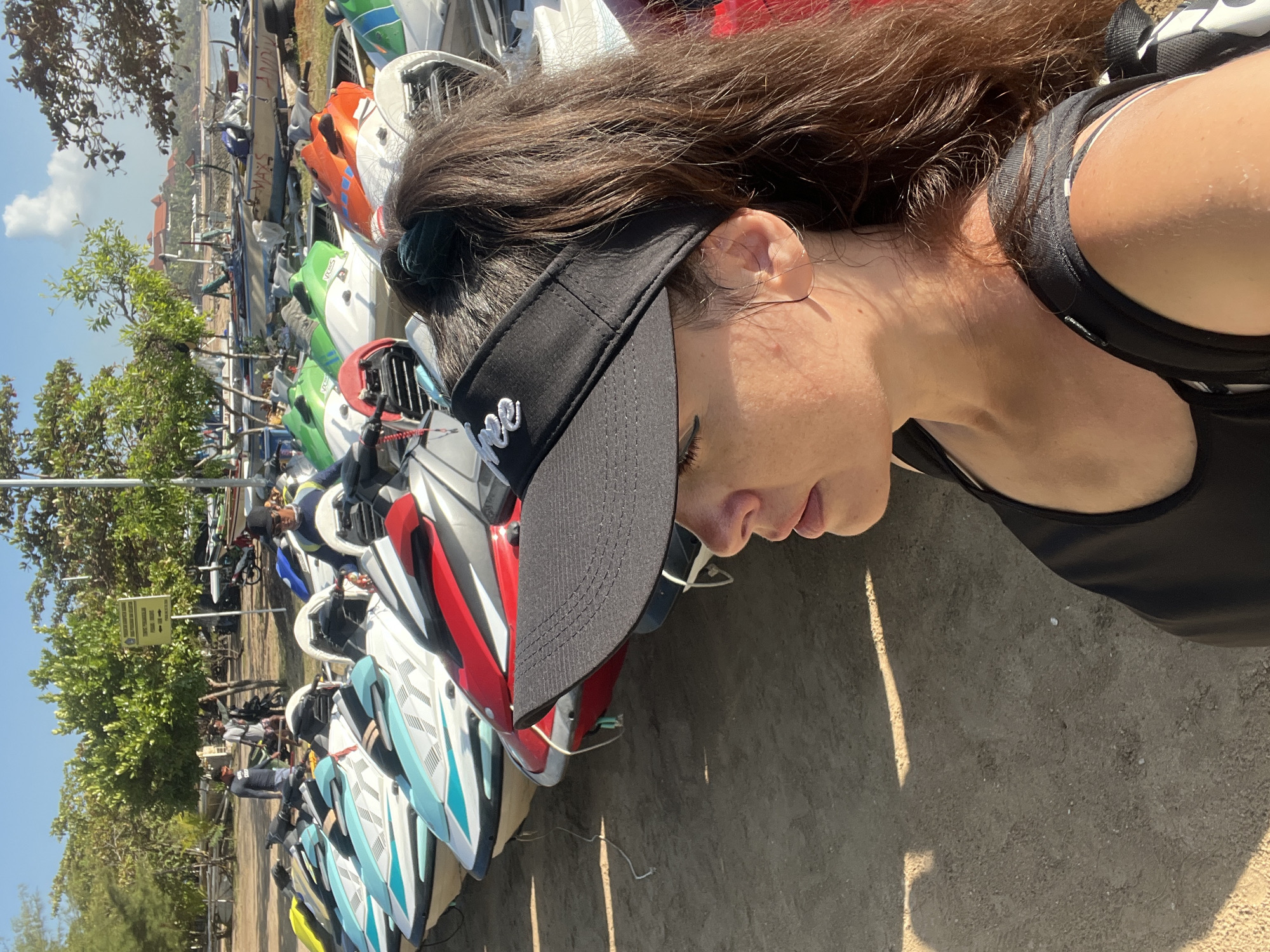 A woman wearing a black visor and a black tank top stands on a sandy beach in Kuta Selatan, Indonesia, with a row of jet skis behind her. The jet skis are of various colors, including white, red, blue, and green, and are parked in a line. In the background, trees and a body of water can be seen. The sky is clear and blue. The woman is looking to her left, with her hair pulled back in a ponytail. The lighting suggests it is daytime.