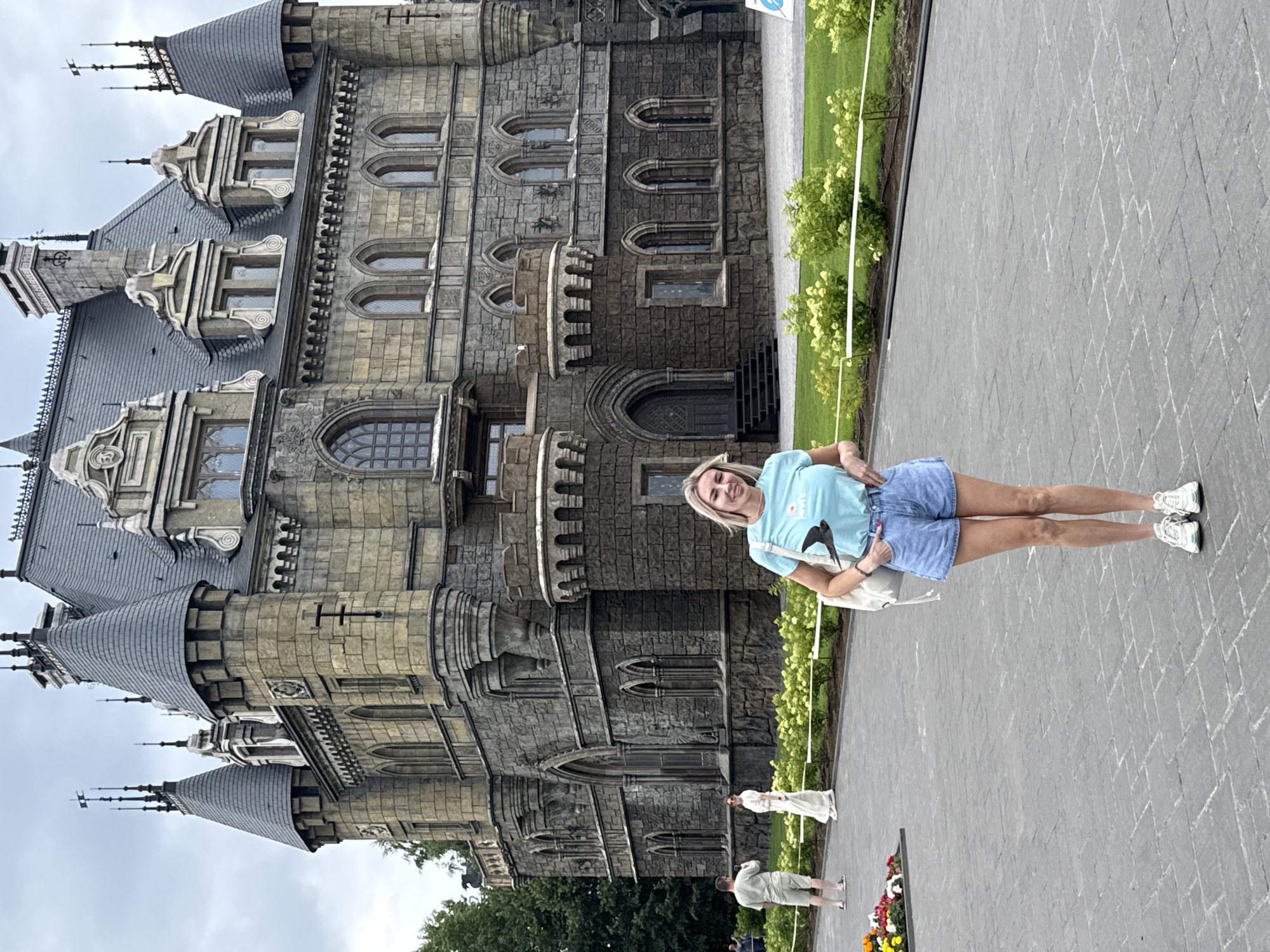 The image shows a woman standing on a paved walkway in front of a stone castle. She is wearing a light blue t-shirt with a graphic of a bird and blue denim shorts. She has a white tote bag slung over her shoulder and is wearing white sneakers. The castle is made of grey stone with a complex architectural style featuring multiple turrets, spires, and arched windows. Green bushes and a lawn are visible to the right of the walkway, and trees are in the background. Two other figures are visible in the distance on the walkway, one wearing a white dress and the other a light-colored shirt and shorts. A flowerbed is also visible in the lower right corner. The sky is overcast, suggesting a cloudy day. The woman is smiling, and the overall mood appears to be relaxed and tourist-like. The text "Khryashchevka, Russia" provides location context.