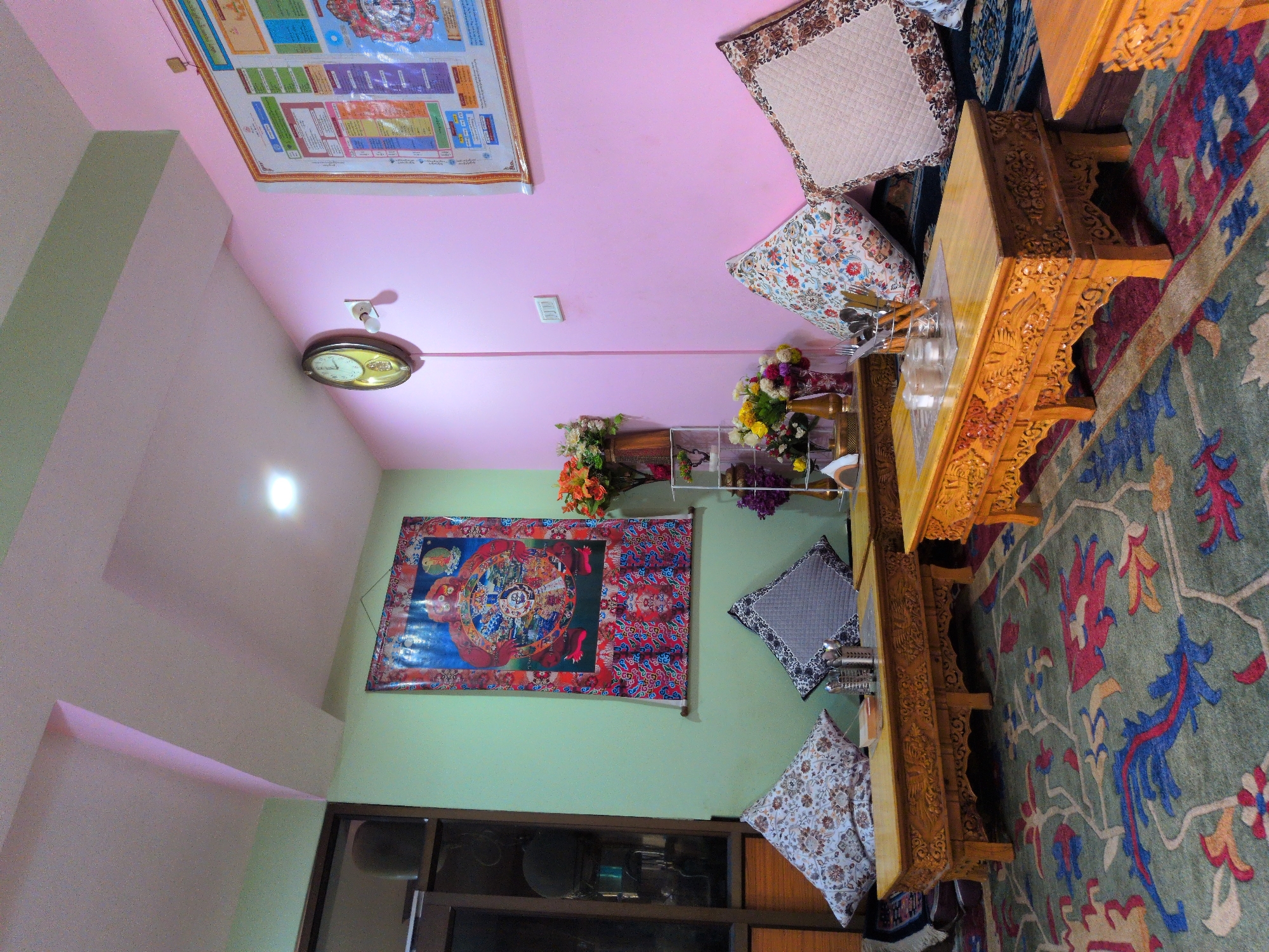 The image depicts an indoor scene, likely a seating area in a cafe or guesthouse in Leh, India, characterized by its Tibetan-influenced decor. The walls are painted in two contrasting colors: a pale green on the left and a soft pink on the right. The ceiling features an interesting architectural detail with a pink accent.
Dominating the left wall is a large, colorful tapestry or thangka, depicting a spiritual or mythological scene. Below this tapestry are two low, intricately carved wooden coffee tables, made of a light-colored wood, possibly walnut or deodar, and adorned with detailed carvings. On the tables, there are cushions with floral and patterned covers, and metal holders containing cutlery.
The right wall is adorned with a framed informational chart or poster with various sections and text. A classic round clock with a golden frame is mounted on this wall, along with a light switch. Two more low wooden coffee tables with similar carvings are also present, with cushions placed on them.
The floor is covered by a large, richly patterned carpet in shades of green, blue, red, and yellow, with intricate floral and vine motifs. In the background, a shelving unit holds vases with artificial flowers in shades of orange, yellow, and purple. A window with dark wooden frames is partially visible on the far left. The overall impression is one of a cozy, culturally rich, and well-decorated space.