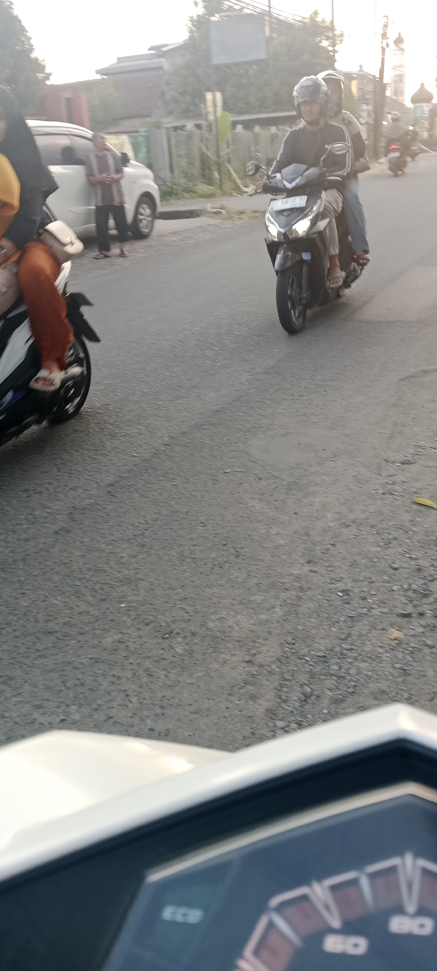 The image shows a street scene in Maguwoharjo, Indonesia, possibly during late afternoon due to the soft lighting. In the foreground, the dashboard of a white scooter is visible, with a digital display showing "ECO" and speed indicators. In the mid-ground, two people are riding a black motorcycle, with the driver wearing a helmet and a dark jacket, and the passenger behind him. They are on a paved road. To the left, a white car is parked, and a person in a brown and white striped shirt and dark pants is standing next to it, leaning against the car. Another person, partially visible, is wearing an orange garment and a black headscarf and is seated on a white scooter that is in motion. The background features a line of trees and some buildings, including what appears to be a minaret of a mosque. The overall environment suggests a residential or commercial area within the city.