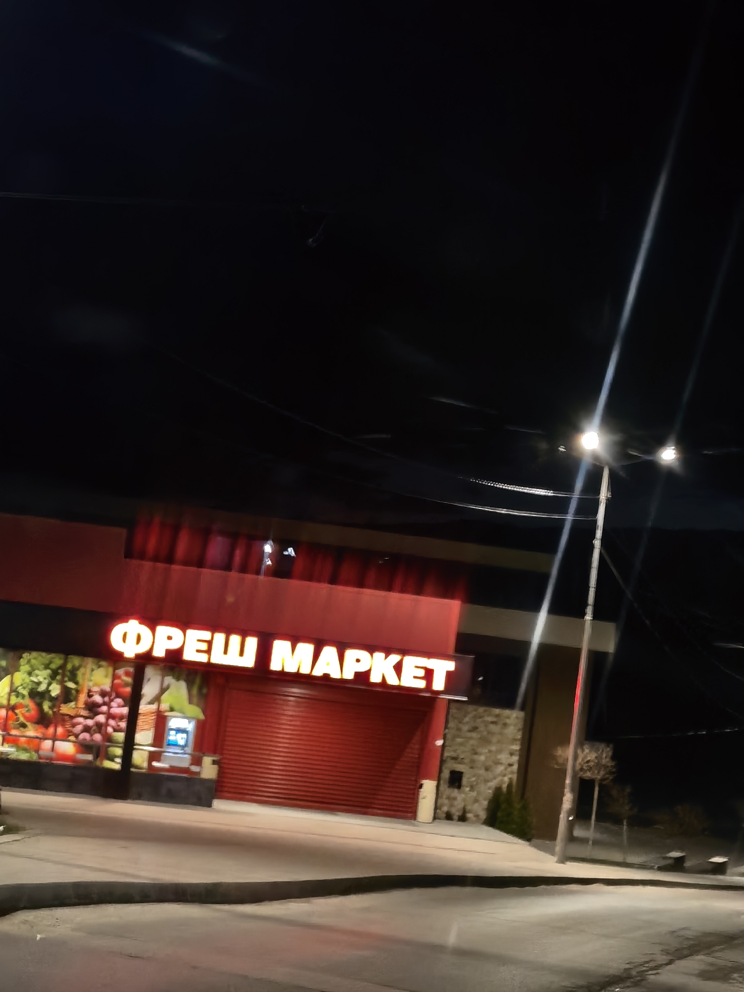 The image shows a "ФРЕШ МАРКЕТ" (FRESH MARKET) store at night in an unidentified city. The building has a red façade with a sign in illuminated yellow letters. The storefront features a display window showcasing fruits and vegetables, and a red roll-up door covers what appears to be the main entrance. An ATM is visible near the store entrance.
A streetlight stands near the store, casting bright illumination. The street is paved, and some vegetation is visible in the background. The sky is dark, suggesting it is late at night. There are no people visible in the scene.