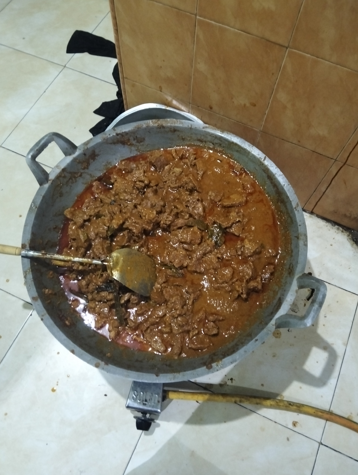 An overhead, medium shot captures a large, well-used, aluminum cooking pot filled with a rich, brown stew, likely beef rendang, a popular Indonesian dish. The stew has a thick, oily sauce with visible pieces of meat. A metal spatula with a wooden handle is partially submerged in the stew, suggesting recent preparation or serving. The pot rests on a portable gas stove, indicated by the "SOLID" brand name visible on its side and a yellow gas hose extending from it. The scene takes place indoors, on a tiled floor with light-colored square tiles. In the background, a tiled wall with brown, square tiles is visible. A piece of black fabric is draped on the floor near the wall. The lighting is dim, casting shadows from the pot and stove. The overall impression is of a home-cooked meal being prepared or served, possibly in a modest setting within the city of Medan, Indonesia, a region known for its diverse culinary heritage.