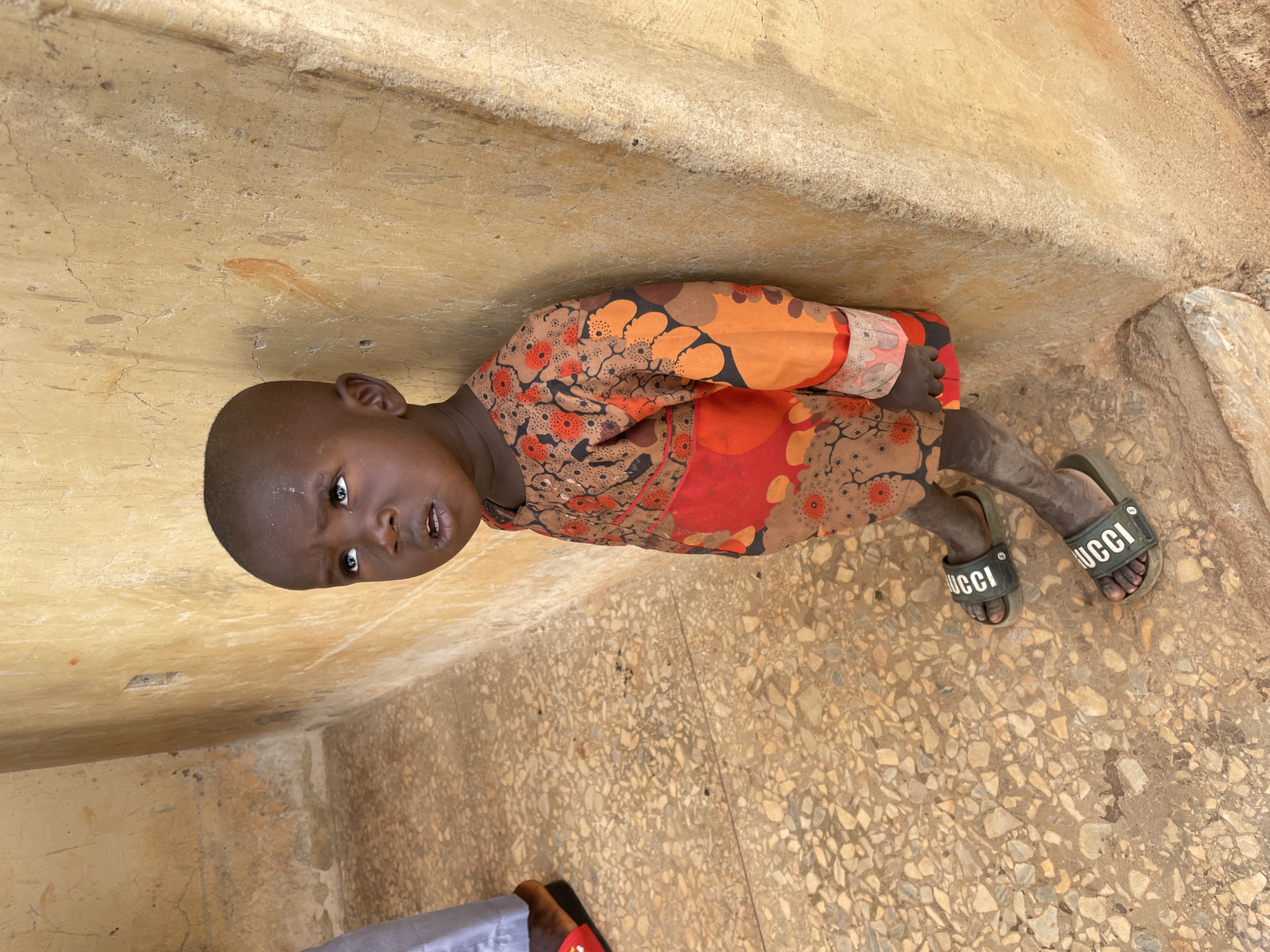 A young child, with dark skin and a close-cropped haircut, stands or sits in a corner formed by two walls. The child wears a long-sleeved shirt adorned with a vibrant, abstract pattern in shades of orange, brown, red, and black. On their feet are dark green slider sandals, each featuring the brand name "GUCCI" in white capital letters. The child looks directly at the camera with a neutral and slightly curious expression.
The walls are made of a light brown, textured material, possibly concrete or an earthen construction, exhibiting cracks and signs of wear. The ground is a light-colored, speckled surface, resembling terrazzo or a mix of small stones. Natural daylight illuminates the scene, suggesting it is daytime. The visual cues of the setting suggest an outdoor or semi-enclosed space in a rural or urban environment. This scene is located in Ladura Foron, Nigeria.