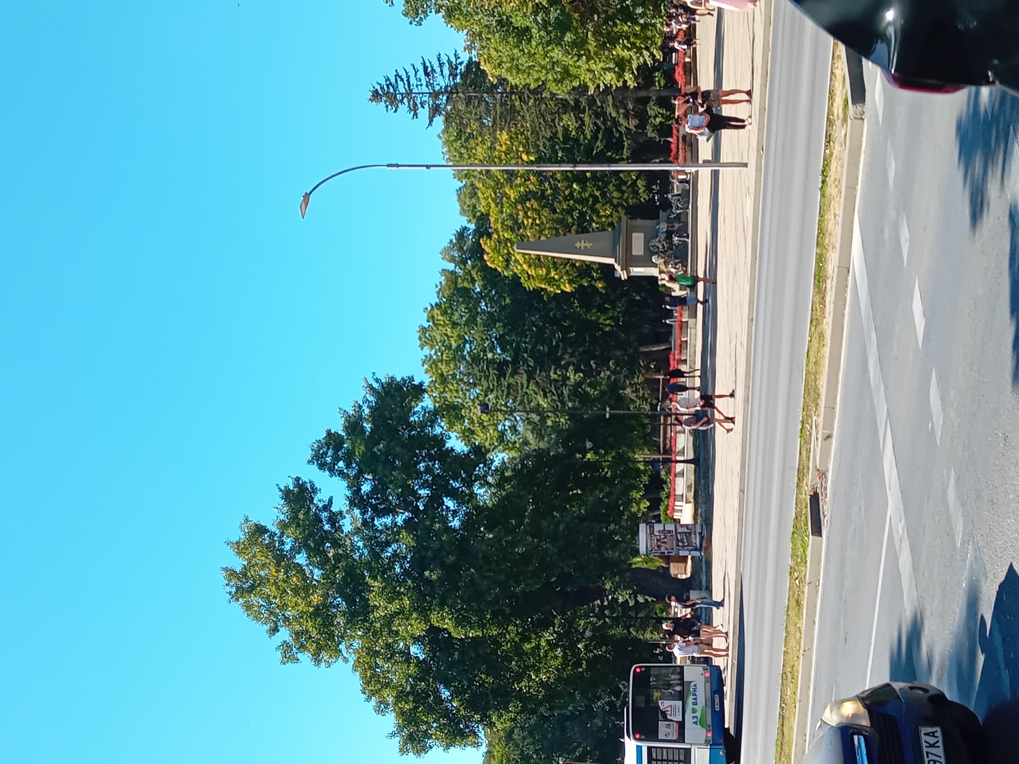 The image captures a sunny day in Varna, Bulgaria, likely in a public square or park area. Dominating the left side is a large, mature tree with lush green foliage, casting shadows. In front of the tree, a white and blue bus is parked, displaying text that includes "АЗ ВАРНА" (meaning "I am Varna" or "From Varna"). Several people are standing near the bus.
Further into the scene, a monument is visible, a tall obelisk-like structure with decorative elements at its base and a symbol at its apex. It appears to be a historical or memorial monument. A street lamp with a curved arm stands to the right of the monument, and more trees form a dense backdrop.
The area in front of the monument seems to be a pedestrian walkway or plaza, with more people walking around. Flower beds with red flowers are visible along the edge of this plaza. A paved road with lane markings runs across the foreground, with a dark-colored car visible on the left side of the road, its headlights on. The sky is a clear, bright blue. The overall impression is that of a vibrant urban public space on a pleasant day.