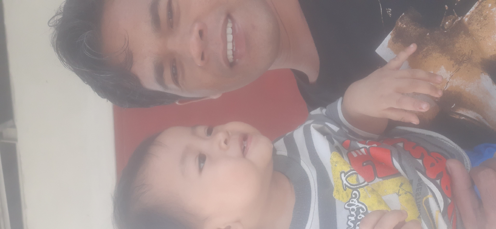 A close-up, hazy selfie features a man and a young child. The man, positioned on the right, has dark hair and is smiling broadly, showing his teeth. He is wearing a black t-shirt with a graphic print on it. The child, on the left, is looking slightly to the side and has a sweet expression. The child is wearing a grey and white striped long-sleeved shirt with a colorful graphic, possibly cartoon characters, on the front. The background is out of focus, but appears to be a building with a red wall and a lighter colored ceiling or overhang. The lighting suggests an outdoor setting, likely during daytime. The image is believed to have been taken in Sepakek, Indonesia.