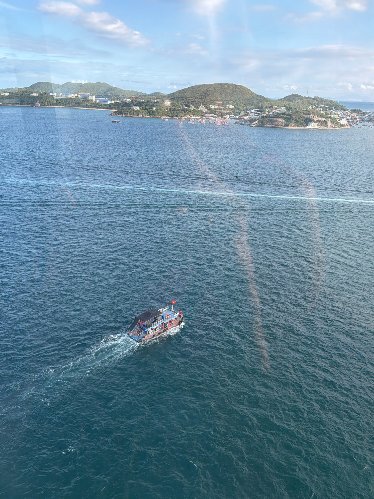 An aerial view captures the turquoise waters of the sea near Nha Trang, Vietnam. A small, colorful boat with a dark roof sails across the water, leaving a white wake. In the distance, a lush, green coastline dotted with buildings and trees stretches across the horizon. The sky above is a mix of blue and white clouds, with sunlight casting a gentle glow on the scene. The image appears to be taken from a high vantage point, possibly a drone or a tall building, offering a panoramic perspective of the coastal landscape.