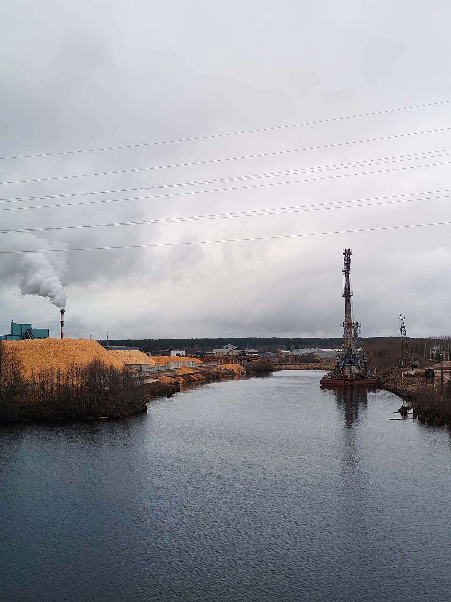 The image depicts a river with industrial elements on both banks under an overcast sky. On the left bank, a large pile of wood chips or sawdust is visible, alongside a factory or industrial building with a prominent chimney emitting a plume of smoke. On the right bank, there are two large cranes, one on a barge in the river. Power lines stretch across the sky. The scene suggests an industrial area, possibly a lumber mill or related facility, along a waterway. The location of Depo, Russia, is not confirmable with this image alone.