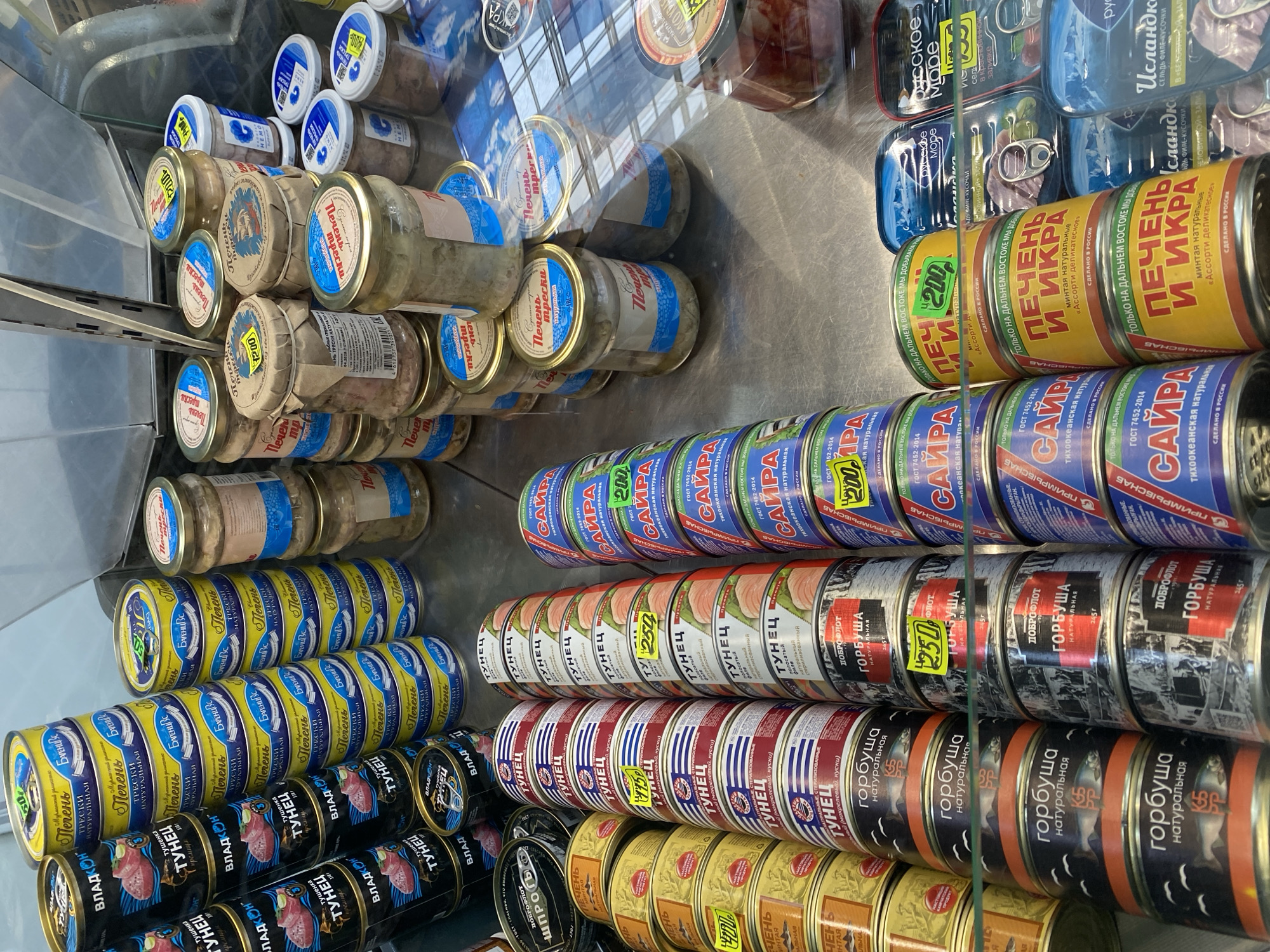 The image shows a retail display of canned fish products, likely in a supermarket or specialty food store in Novaya Usman', Russia. The products are arranged on shelves, with the majority being cans of various sizes, and some glass jars also visible. The lighting suggests it is daytime, possibly indoors under artificial store lighting.
The cans and jars are densely packed, creating multiple rows and layers of products. Many of the labels are visible and feature Cyrillic text. Some of the prominent text visible on the cans includes "САЙРА", "ТУНЕЦ", "ГОРБУША", and "ПЕЧЕНЬ И ИКРА". There are also price tags, with numbers like "200" and "250" visible. The glass jars contain what appears to be preserved fish or seafood, with labels indicating "Печень трески".
The overall scene is one of commercial inventory, with a focus on a variety of preserved seafood items. The reflections on the glass in front of the display suggest it's a refrigerated unit. There are no people visible in the image, and the focus is entirely on the product arrangement. The colors of the packaging vary from bright yellow and blue to darker, more muted tones.