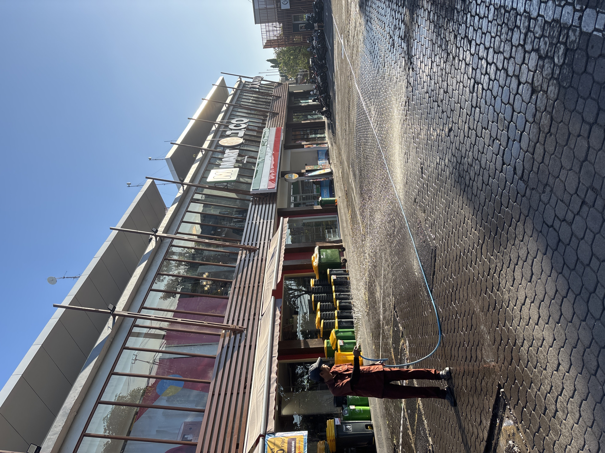 A person is cleaning a brick pavement area in front of a building in Kuta Selatan, Indonesia. The person is wearing a dark hat, a dark red long-sleeve shirt, and dark red pants. They are holding a blue hose and spraying water onto the pavement. To the side are rows of yellow, green, and black bins. The building behind the person is multi-story and has the signage "K.MART 100" visible. There are motorcycles parked in front of the building to the right. The sky is clear and blue.