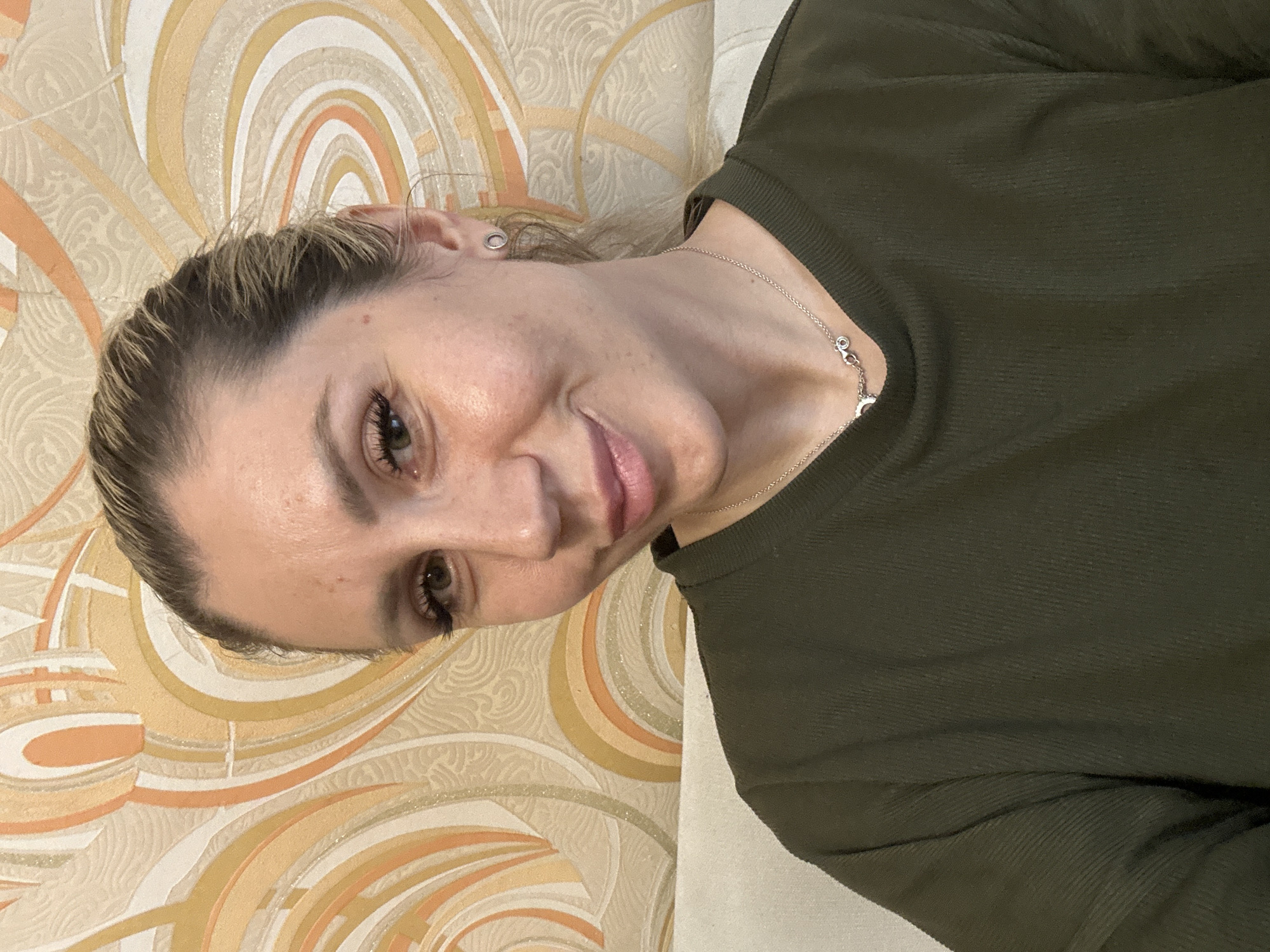 An adult Caucasian female with blonde hair pulled back is shown from the chest up. She is looking directly at the camera with a slight smile. She is wearing a dark green long-sleeved shirt and a silver necklace with a small pendant. She has a small silver earring in her right ear. The background is a patterned wallpaper in shades of beige, orange, and yellow. A portion of a light-colored sofa is visible in the lower left of the frame. The scene appears to be an indoor setting, possibly a living room or common area. There is no visible indication of the specific location within Novoaltaysk, Russia.