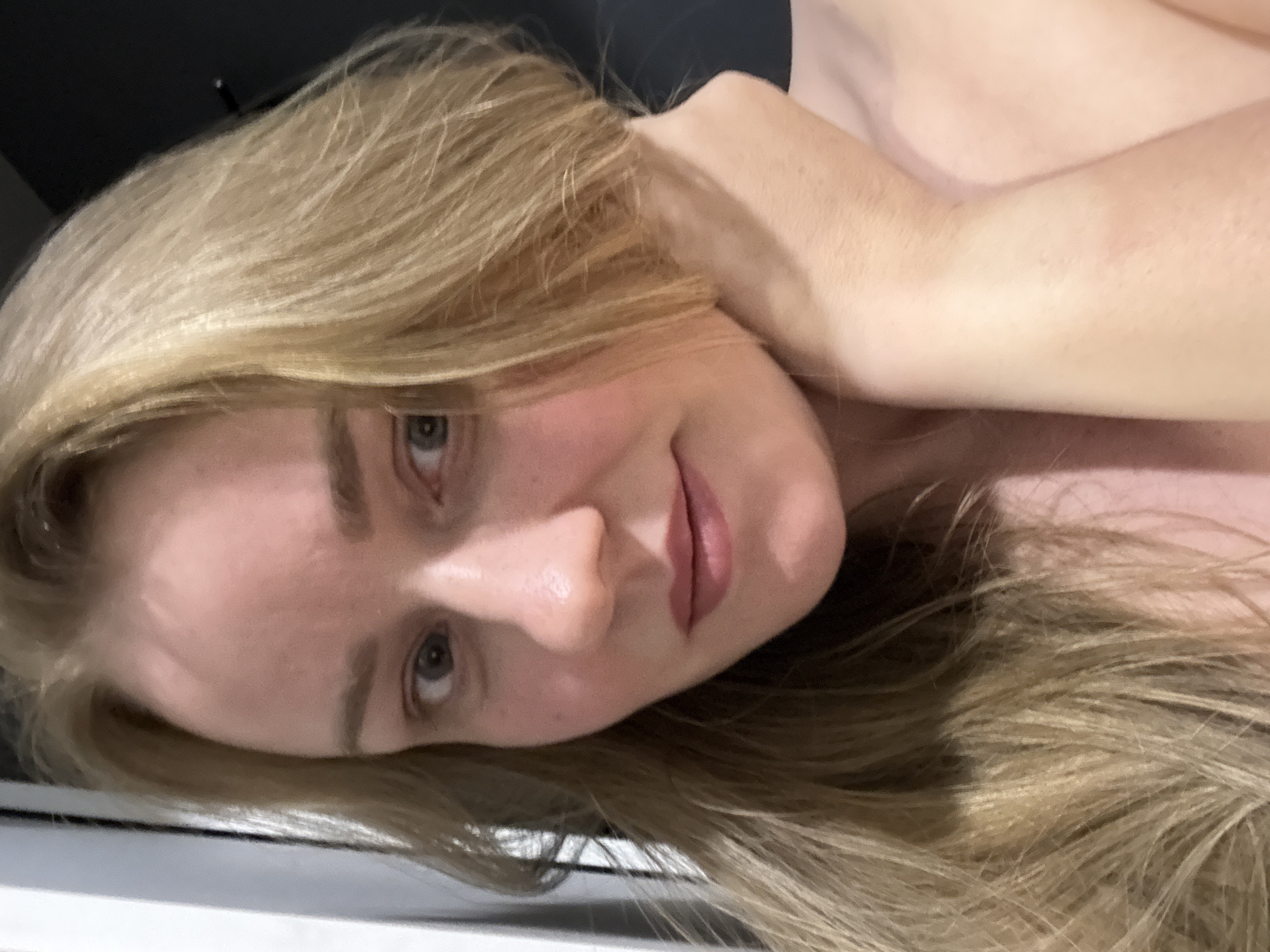A close-up, selfie-style image of a woman with light blonde hair. Her head is resting on her arm. Her eyes are light-colored, and she has a slight smile. The background is dark and out of focus. The image does not contain any information about the location. It is impossible to identify that it was taken in the city of Chaikovsky, Russia.