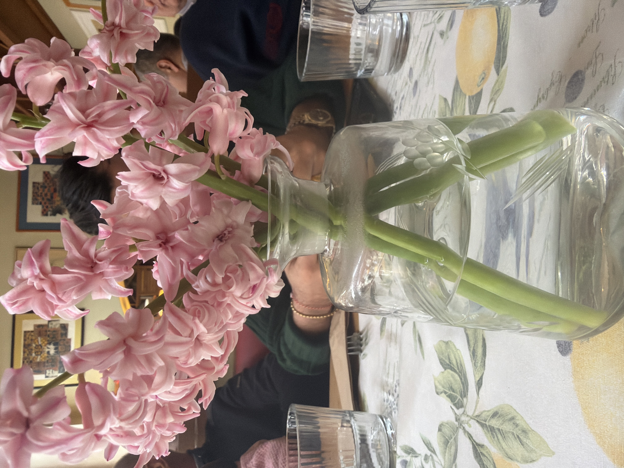 A table setting features a bouquet of pink hyacinths in a clear glass vase filled with water. The stems of the flowers are visible through the vase. Two clear drinking glasses, also filled with water, are placed on the table. The tablecloth has a white background with a pattern of lemons and leaves. People are seated at the table, partially visible in the background; one wears a dark blue garment and another a green garment with gold bracelet. The setting appears to be indoors. Background objects also include framed artwork hanging on the wall, indicative of a domestic or restaurant setting. There is no specific information in the image to establish the location as Sofia, Bulgaria.