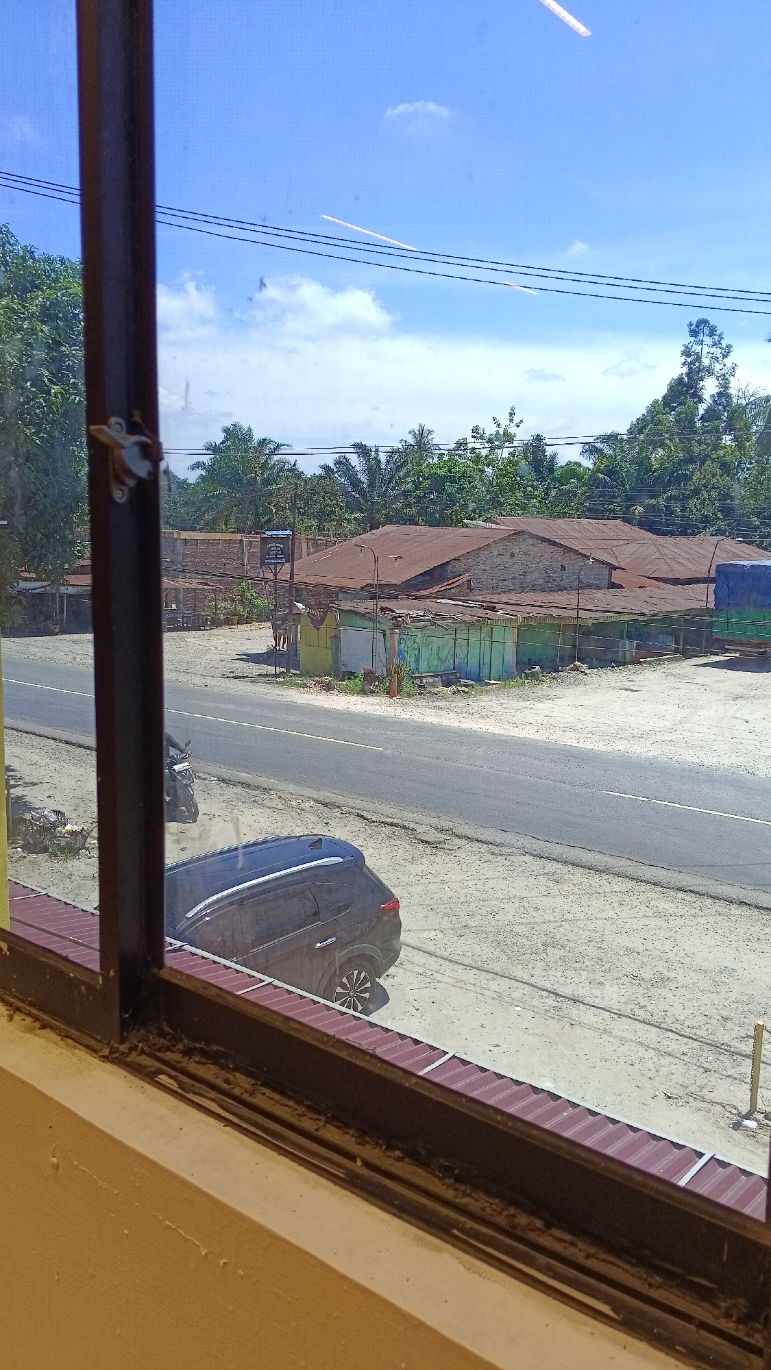 A view through a window of a street scene in Gunting Saga, Indonesia. The foreground shows the bottom of the window frame and the interior wall of the room from which the photo is taken. Outside, a paved road runs horizontally across the image, bordered by a sandy shoulder. On the road, a dark-colored SUV is parked on the right side, facing away from the camera. Further down the road, a motorcycle is visible. Across the road, there are several low-rise buildings with corrugated metal roofs. Some of these buildings appear to be shops or stalls, with one painted in green and light blue. Beyond these structures, palm trees and other greenery are abundant. The sky above is clear blue with scattered white clouds. Two white contrails from aircraft are visible in the sky. Power lines are strung across the upper portion of the scene.