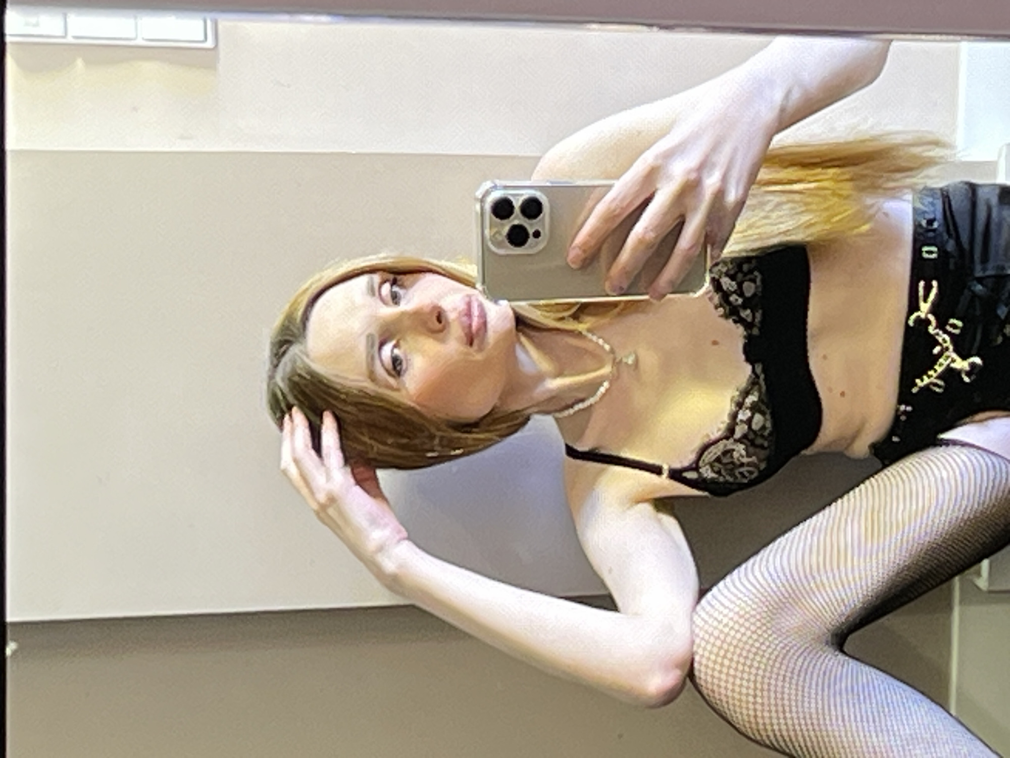 A young woman with long, blonde hair is sitting on the floor, taking a mirror selfie. She is wearing a black lace bra, fishnet stockings, and a black skirt with a gold chain belt. Her left arm is resting on her knee, and her right hand is holding a smartphone. The background is a plain, light-colored wall, and the lighting appears to be artificial. The woman's expression is neutral.