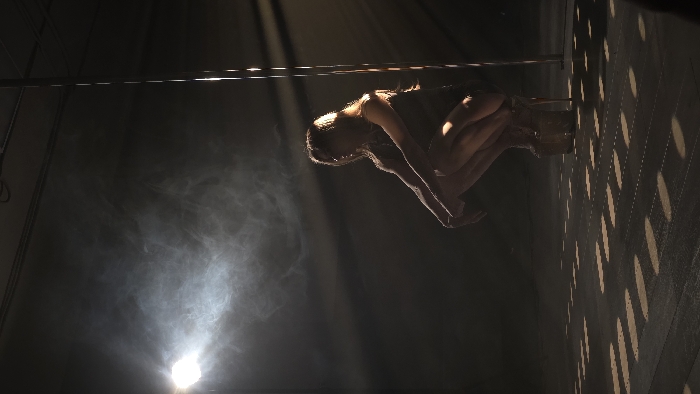 An indoor scene depicts a single female individual in a dimly lit, hazy environment. The woman is positioned in a low crouch or squat, facing slightly right with her head turned to her left. She wears a dark, form-fitting garment and clear, high-platform heels. A strong spotlight from the upper left illuminates the scene from behind the subject, casting her primarily in silhouette and making visible a significant amount of airborne haze or smoke. To the right of the woman, a vertical, reflective metallic pole extends upwards from the floor. The floor beneath the woman displays a distinct pattern of illuminated parallel dashed lines. The specific location within Krasnoyarsk, Russia, is not identifiable from the visual information presented.