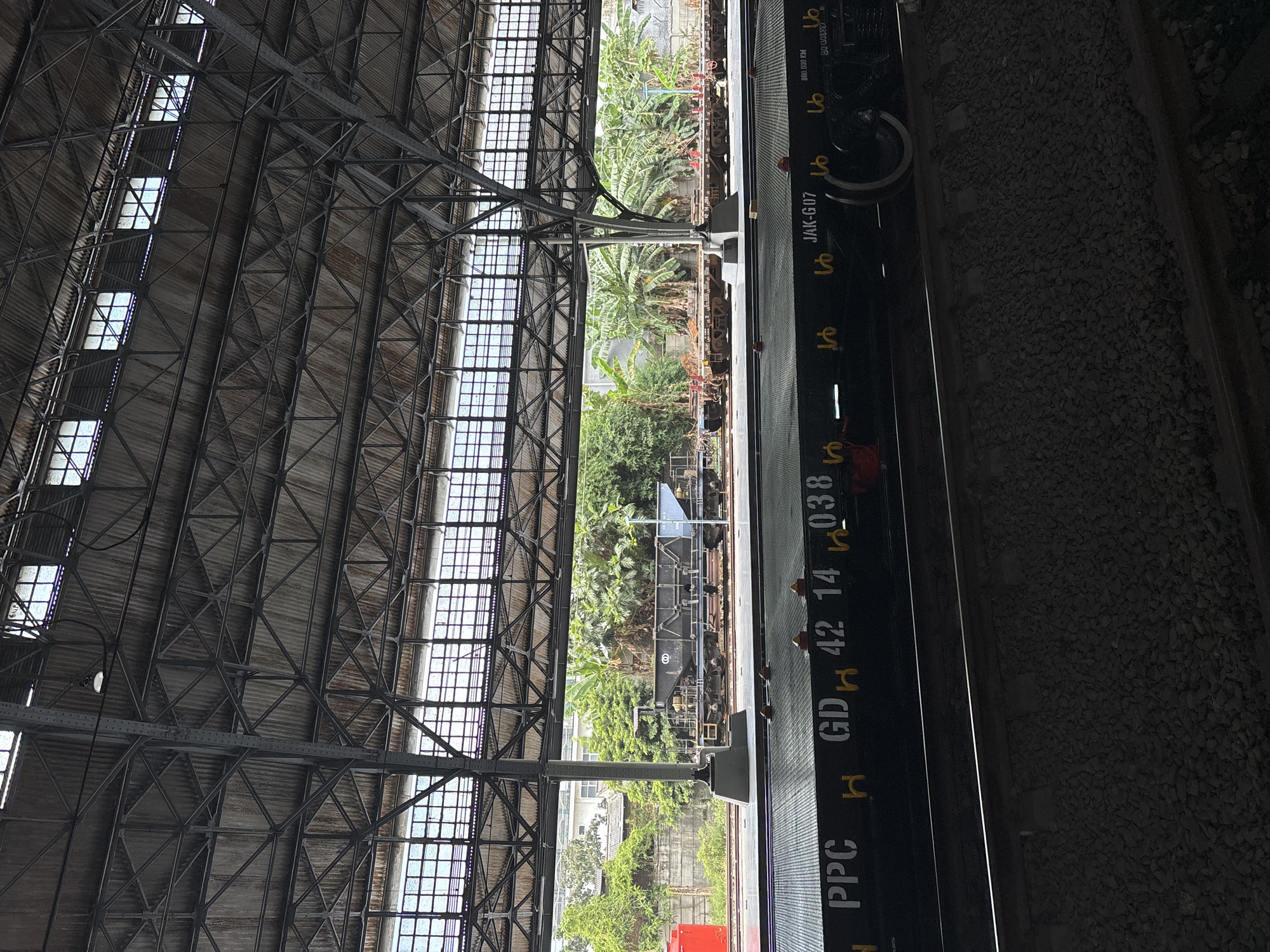 The image captures an expansive view inside a large, industrial-style railway station or train shed, likely in North Jakarta, Indonesia, during daylight hours. The dominant feature is the massive roof structure, composed of intricate metal trusses and corrugated panels, interspersed with numerous rectangular skylight windows that allow bright sunlight to filter in.
Below the roof, a concrete platform runs alongside a set of railway tracks. On the tracks, a dark-colored train car or freight wagon is partially visible, primarily on the right side of the image. Lush, tropical greenery, including prominent banana trees and other leafy vegetation, fills the open space beyond the platform, suggesting an outdoor area adjacent to the covered structure. In the far distance, parts of residential buildings can be seen through the foliage.
No people are visible in the frame. The scene is static, devoid of active movement, conveying a sense of quiet waiting or storage.
Visible text and numbers on the side of the train car or platform edge include: "PPC", "GDH 42 14 038", "JAK-G07", "800.000 KM", and "BO-1308887". Additionally, several yellow stylized symbols, resembling either cursive 'Lo' or the Arabic letter 'ma' (ما), are repeated along the dark surface.