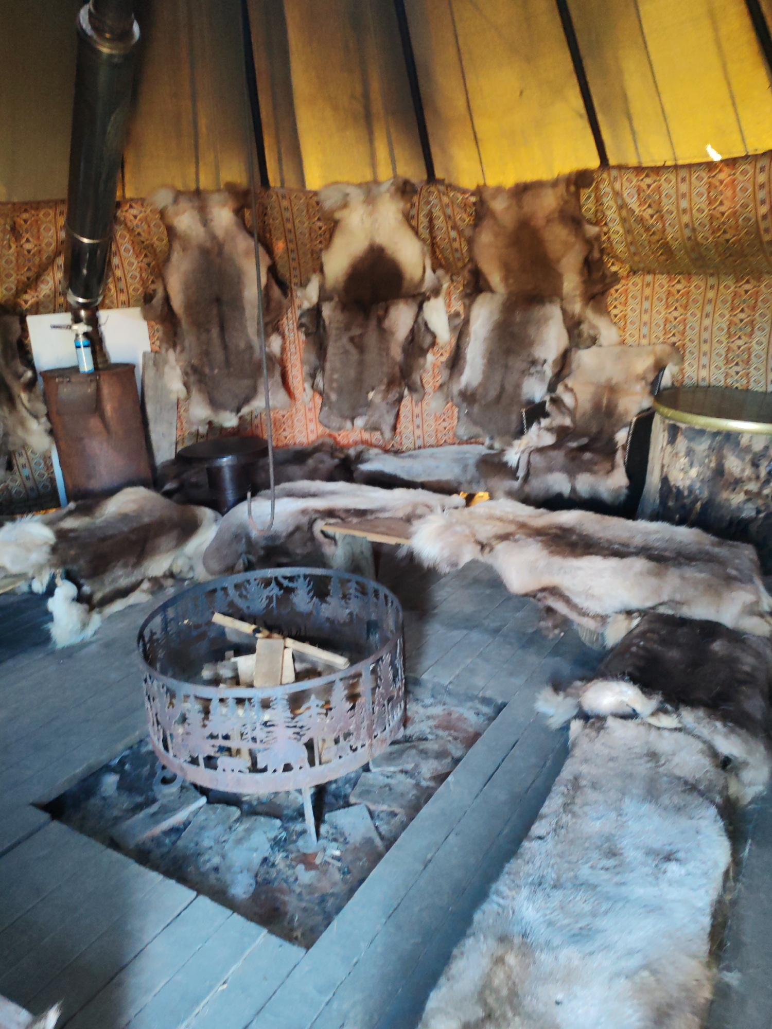 The image depicts the interior of a dwelling, likely a traditional tent or yurt, with wooden floors and fabric walls. Various animal pelts are draped over furniture and hung on the walls, suggesting a rustic or indigenous setting. A metal fire pit, containing firewood, is positioned on the floor in the foreground. A tall metal pipe, possibly a stovepipe, extends upwards on the left side of the frame. The overall impression is one of a lived-in, primitive shelter. There are no people or specific identifiable landmarks to ascertain a precise location within Lukaši, Russia, beyond it being an interior space.