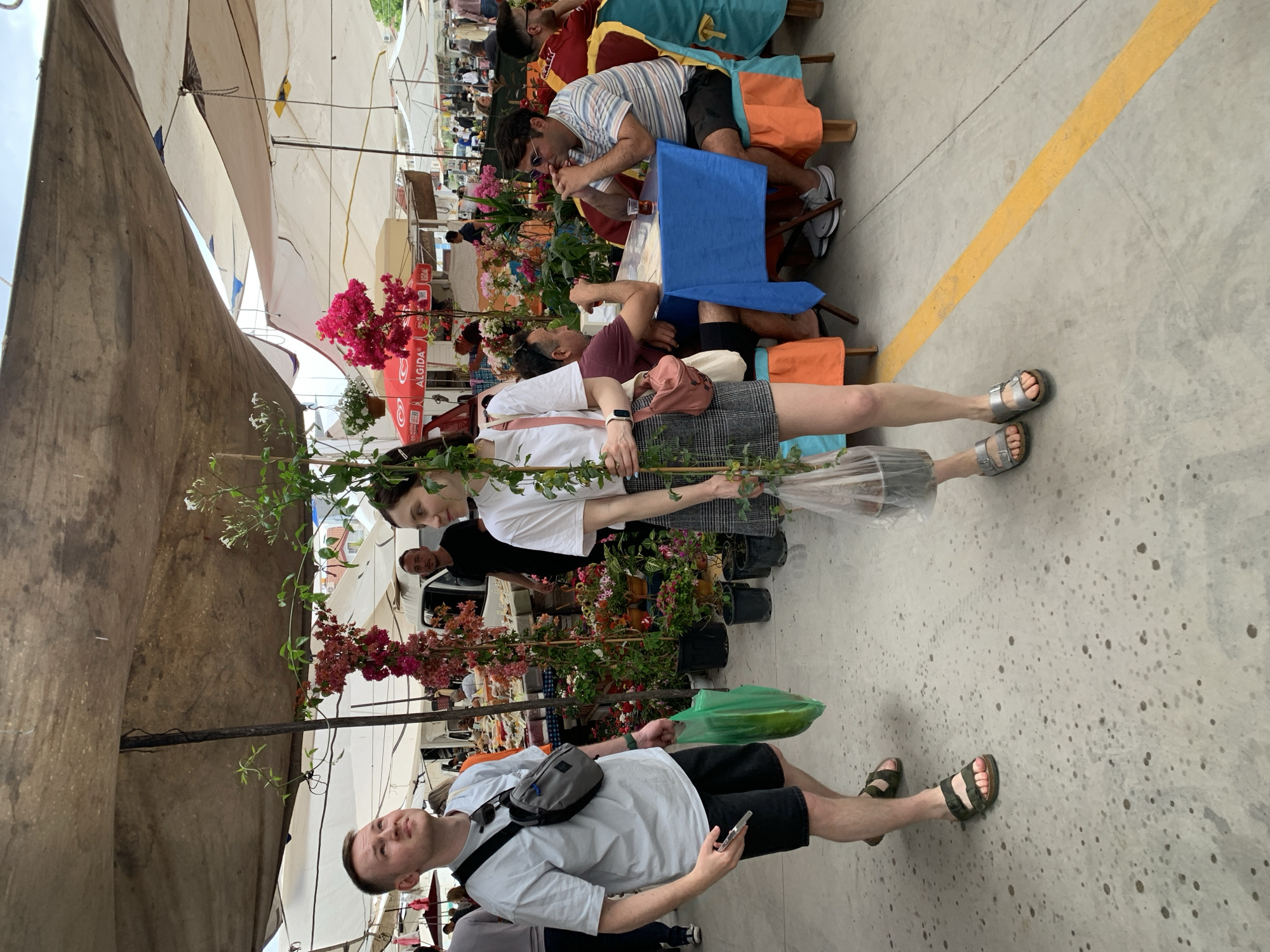 The image shows a scene at a market in Fethiye, Turkey. Two people, a man and a woman, are standing in the foreground, holding small potted plants with flowers and green foliage. The man is wearing a light-colored t-shirt, black shorts, and sandals, with a small crossbody bag. He is holding a smartphone in his hand. The woman is wearing a white t-shirt, a checkered skirt, and silver sandals. Behind them is a row of potted plants and flowers for sale. Further back, there are people sitting at tables under awnings, presumably eating or drinking. The floor is concrete with a yellow line painted on it. The scene is outdoors, with market stalls and awnings providing shade.