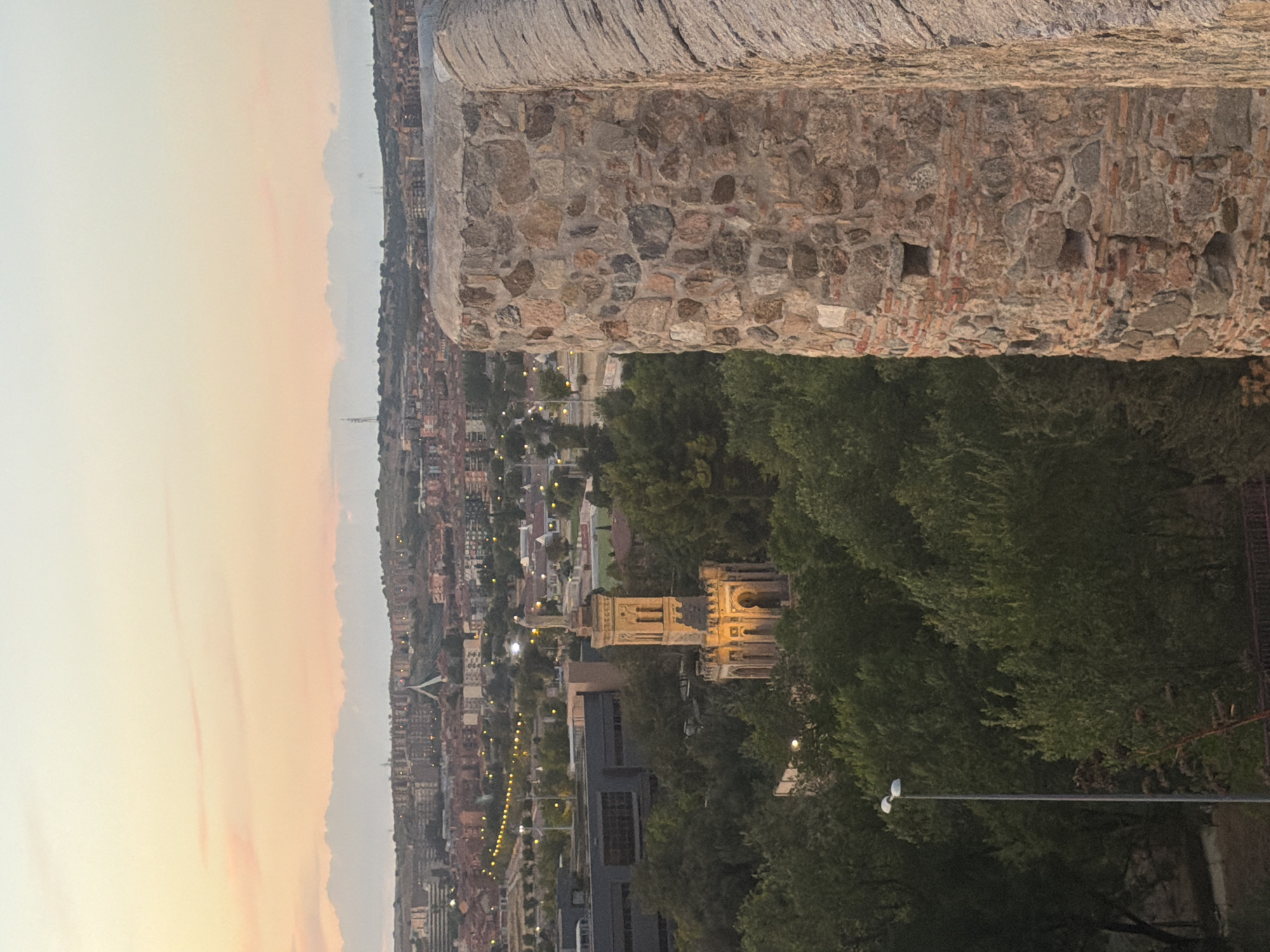 This is a medium shot, taken from an elevated vantage point, capturing a cityscape of Toledo, Spain at dusk. In the foreground on the right, a section of a thick, ancient stone wall is visible, constructed from rough-hewn, varied colored stones. Its weathered texture suggests considerable age. To the left of the wall, a verdant expanse of trees fills the mid-ground, obscuring much of the city below.
Prominently featured within the trees is a detailed, ornate tower. The tower is illuminated from below with warm, golden light, highlighting its architectural features: arched windows, decorative stonework, and a statue of a figure standing atop it. Adjacent to this tower, a modern, dark-colored building with geometric windows is partially visible.
Further in the background, the sprawling cityscape of Toledo unfolds. Buildings with terracotta roofs are densely packed, interspersed with streetlights that cast a warm glow across the urban landscape. The distant horizon is marked by a gently sloping hill, on which more buildings are scattered. The sky above is a soft gradient of pale orange and pink, indicating the final moments of sunset, with a few wispy clouds adding texture to the scene. A tall, slender lamppost with two modern light fixtures stands on the left side of the frame, near the bottom edge. The overall impression is a blend of historic architecture and modern urban development set against a tranquil, twilight sky.