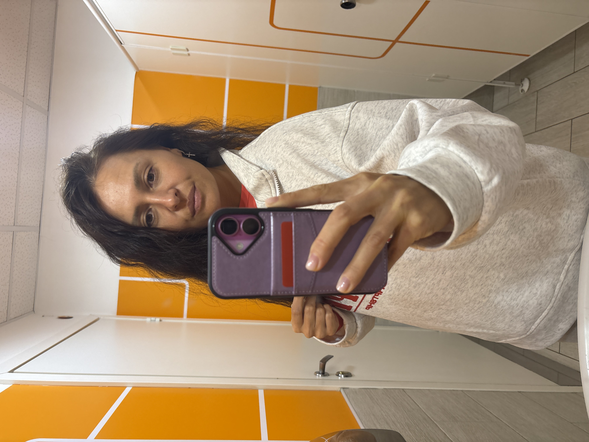 A woman takes a mirror selfie indoors, holding a smartphone in a purple case. She is centered in the frame, looking directly into the camera with a neutral to slightly smiling expression. Her dark, long hair falls over her shoulders. She is wearing a light grey speckled (heathered) zip-up sweatshirt over a red top. A small cross earring is visible on her left ear (viewer's right).
The setting appears to be an indoor facility in Можга, Russia, likely a changing room, locker room, or a well-maintained public restroom. The background features white walls and cabinetry accented with bright orange panels, creating a clean and modern aesthetic. A white tiled ceiling with grid lines is visible at the top of the frame, and a grey tiled floor is seen at the bottom. A silver door handle or latch is visible on a white door in the lower right portion of the reflected image.
The smartphone case is purple with a prominent red stripe or card slot visible on the back. Partially visible on her red undershirt, through the reflection, is mirrored Cyrillic text that appears to read "ЧНЫЙ БЛАН". The image captures an everyday moment in a brightly lit, functional indoor environment.