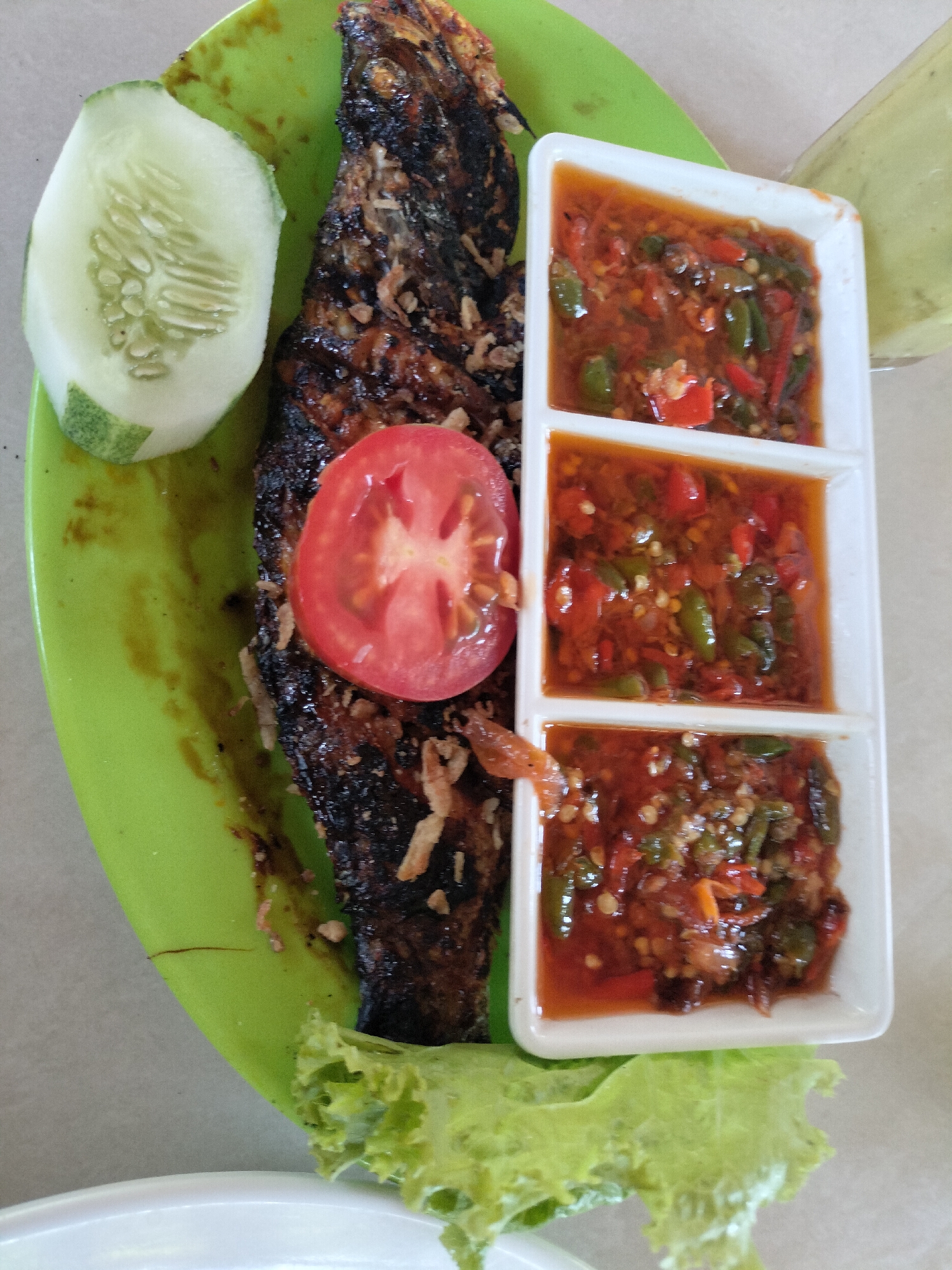 The image shows a plate of food, likely a meal served at a restaurant or home in Tambonbaroh, Indonesia. The main subject is a whole grilled fish, dark brown and slightly charred, placed on a bright green plate. To the left of the fish is a slice of cucumber, and a slice of tomato is positioned on top of the fish. Adjacent to the fish, to the right, is a white condiment dish divided into three sections, each filled with a red chili sauce, possibly sambal. A garnish of lettuce is visible at the bottom of the plate. The background suggests an indoor setting with a light-colored surface, possibly a table. The lighting is bright, indicating daytime.