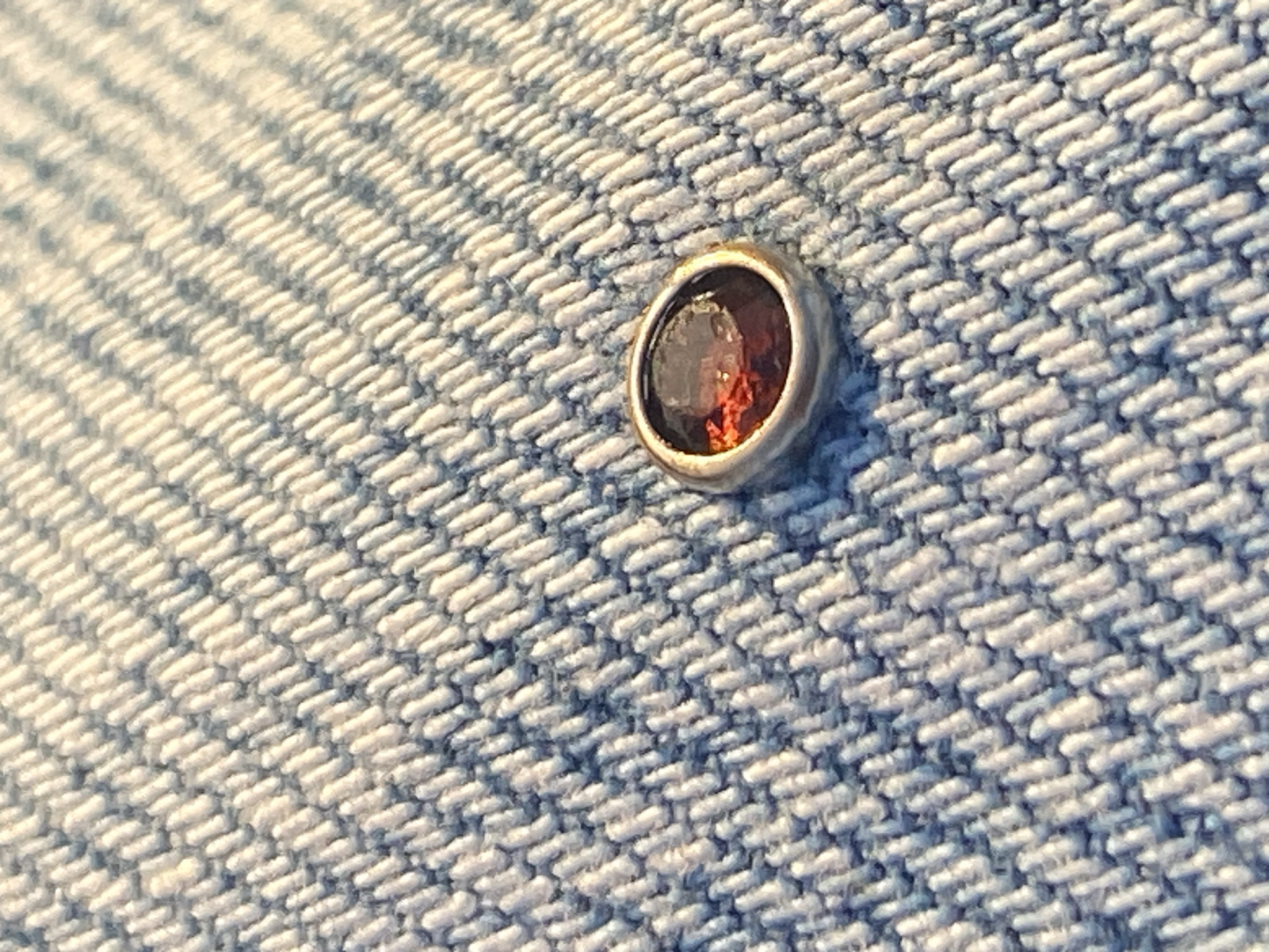 A close-up shot shows a single oval-shaped cabochon garnet set in a silver bezel setting, affixed to a piece of denim fabric. The garnet is a deep reddish-brown color, and the silver bezel has a slight tarnish. The denim fabric is light blue with a prominent woven texture. The image does not provide any information about Tel-Aviv, Israel.