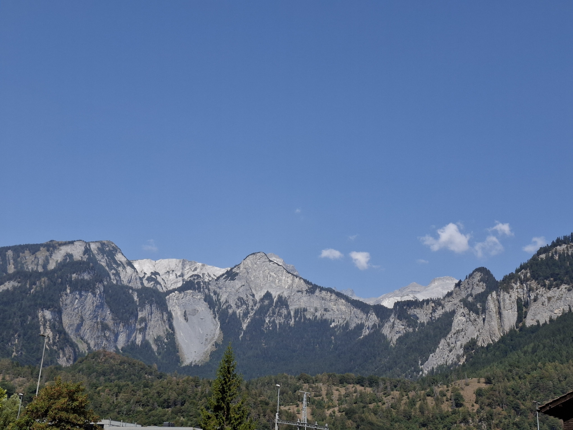 The image depicts a wide, panoramic view of the Swiss Alps near Domat/Ems, Switzerland. The sky is a clear, bright blue with a few wispy white clouds. The foreground features a dense forest of green trees covering rolling hills. In the mid-ground, the imposing, rocky slopes of the mountains rise sharply, revealing stark grey rock faces interspersed with dark green coniferous trees clinging to their sides. Some areas of the mountains show evidence of scree slopes or rock slides. Several tall, thin streetlights are visible in the lower portion of the image, along with some man-made structures, likely part of the infrastructure of the town. The overall scene is one of natural grandeur, with the raw beauty of the mountains dominating the landscape.
