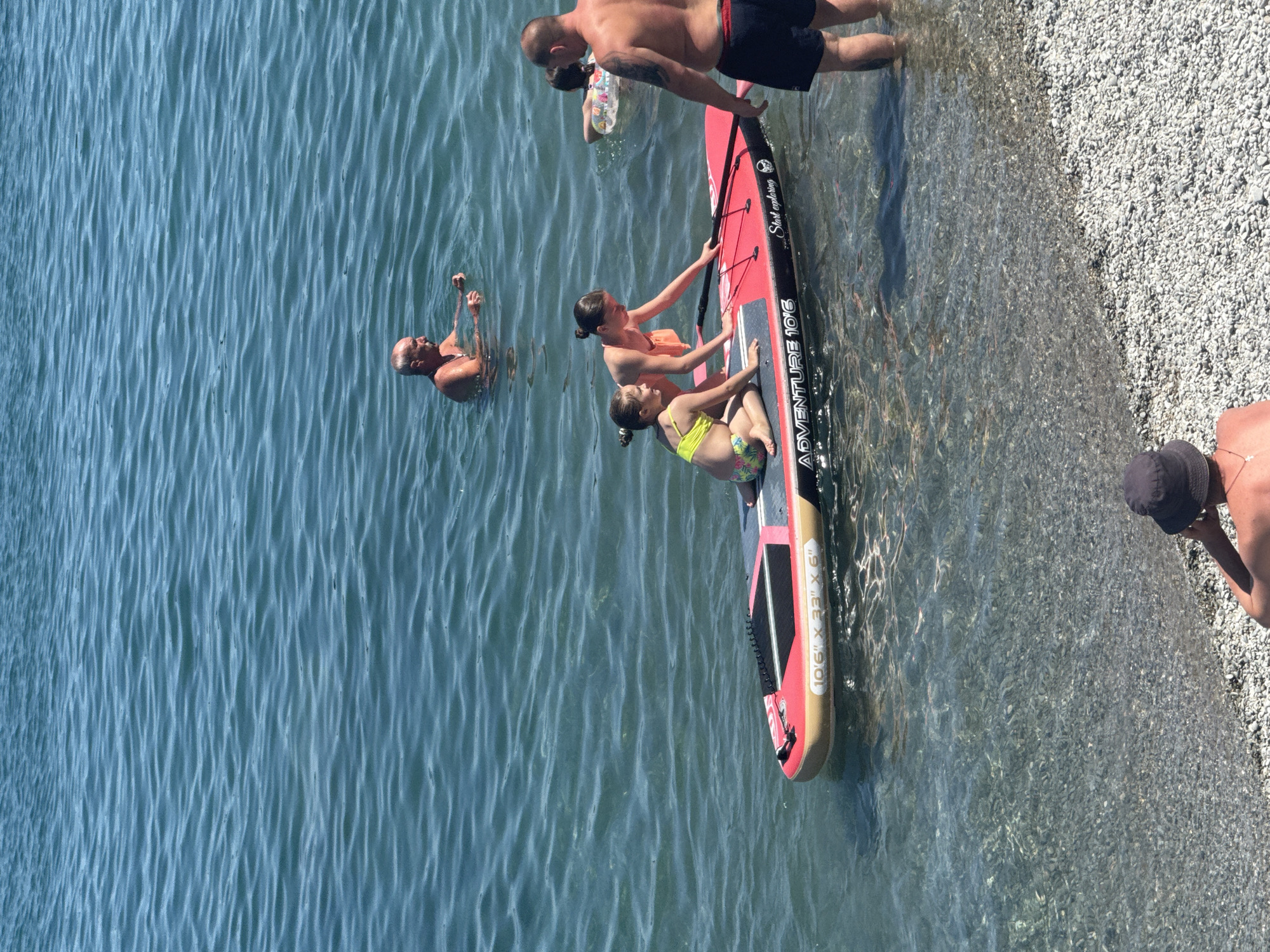 The image shows a family enjoying a sunny day at the beach. The scene is set on the clear, shallow waters of the sea with a pebble beach bordering it. The water is a vibrant blue with small ripples, indicating calm weather.
There are several people in the water and on a paddleboard. In the foreground, two young girls are sitting on a red paddleboard. One girl, wearing a yellow bikini, is looking towards the camera, while the other, in an orange bikini, is reaching out to touch the paddleboard. Behind them, a man in black shorts is partially submerged in the water, leaning on the paddleboard. To the left, another man is also in the water, with his hands raised.
Further back, a person is seen floating in the water with an inflatable ring. On the beach, a person wearing a dark hat is visible in the lower right corner, seemingly looking at their phone.
The paddleboard has text on it, including "ADVENTURE 10'6" and "START EXPLORING". The overall mood is relaxed and joyful, suggesting a pleasant family outing during daylight hours. The pebbles on the beach and the clear water are distinctive features of the location, which is identified as Partenit, Ukraine.