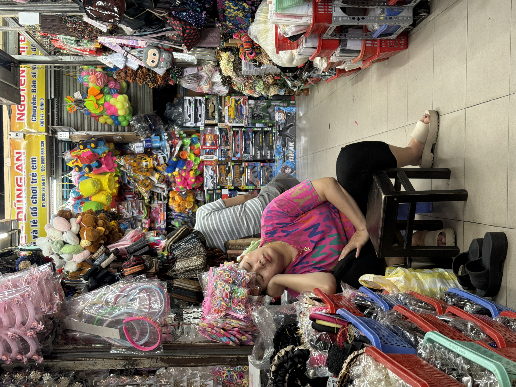 An eye-level, indoor shot depicts a person sleeping amidst a display of goods for sale in a market, potentially in Đà Nẵng, Vietnam. A person is lying on their side atop merchandise, their head resting on packaged hair accessories. They wear a pink patterned shirt and black pants. Another person is partially visible nearby, wearing a striped shirt. A black stool is positioned near the person's legs, with one foot elevated on it. Additional footwear, including white platform sandals and black slides, are visible near the stool. The surrounding shelves are densely packed with a variety of goods, including toys, plush items, hair accessories, bags, and packaged items. A yellow sign with Vietnamese text is also visible. Rows of plastic baskets filled with merchandise are in the foreground. The floor is tiled in a light color.