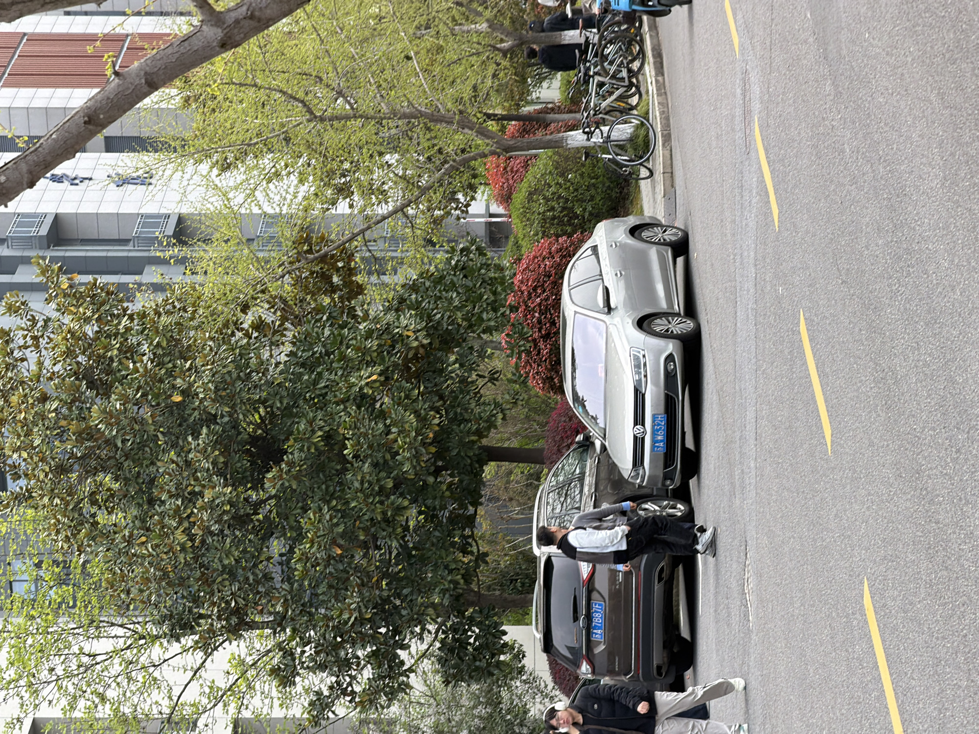 A street scene in Jiangning District, China, shows two vehicles parked on the side of a road, adjacent to a row of shrubs and trees. The first car is a dark-colored SUV with license plate 苏A·7887F. A man stands between the cars, appearing to engage in conversation. A second individual, wearing headphones and a jacket, stands partially visible towards the rear of the SUV. To the side of the SUV is a light-colored sedan, license plate 苏A·N632H. Bicycles are parked near a building at the upper edge of the image. The road has yellow line markings.