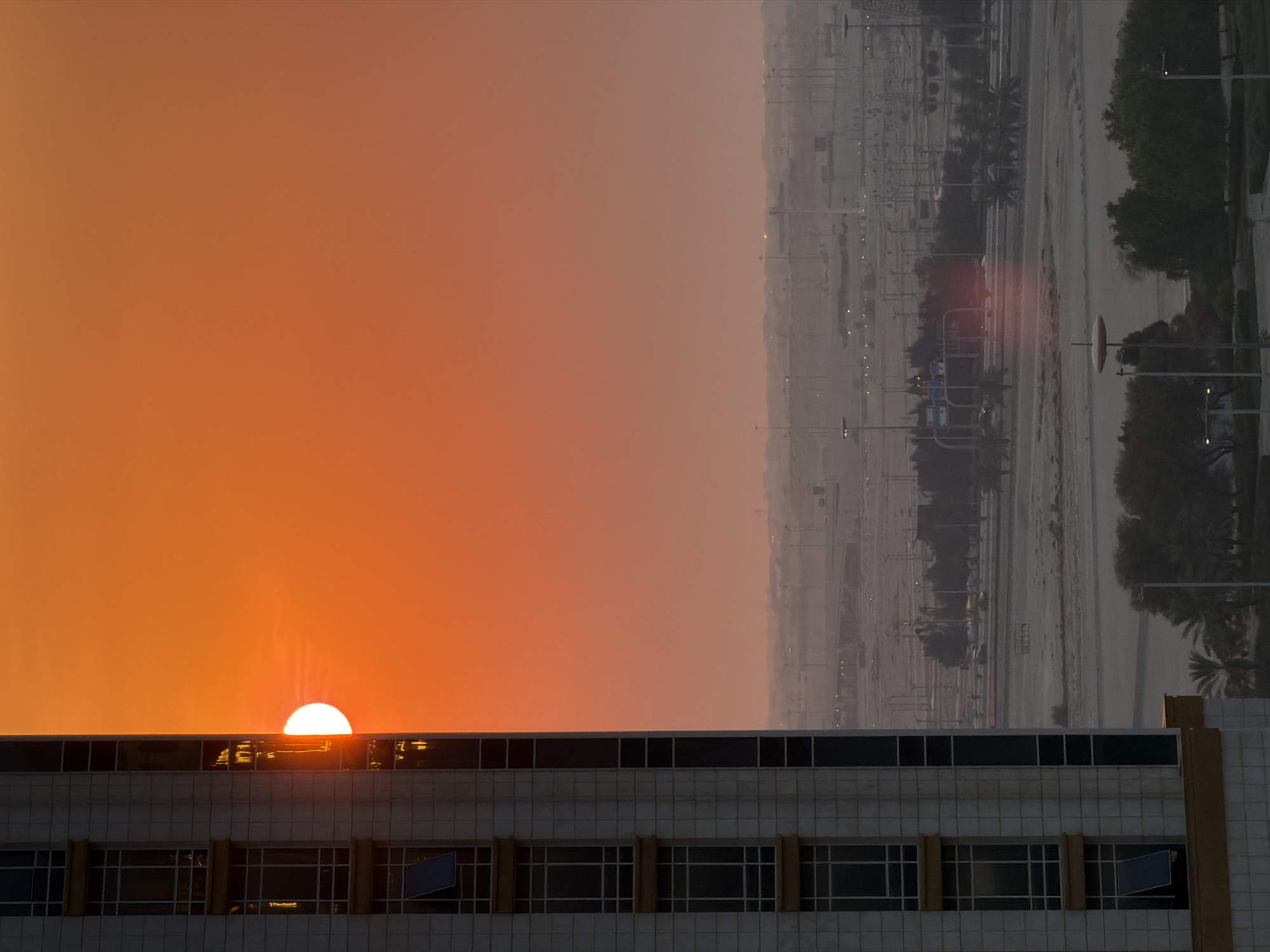 A high-rise building with a light-colored facade dominates the left side of the frame, its windows reflecting the intense orange glow of a setting sun. The sun, a bright disk partially obscured by the building, is positioned on the right side of the building's reflection. The sky is a gradient of warm oranges and reds, fading to a lighter orange towards the horizon. In the middle ground, a vast, flat landscape stretches out, featuring numerous lamp posts and what appear to be roads or pathways, suggesting an urban or suburban environment. Sparse trees are visible, some forming small clusters. The distant horizon is hazy, with hints of structures and activity blurred by the atmospheric conditions. The overall scene is captured during dusk or sunset, highlighting the warm, vibrant colors of the sky and the stark silhouette of the urban landscape. This scene is located in Lusail, Qatar.