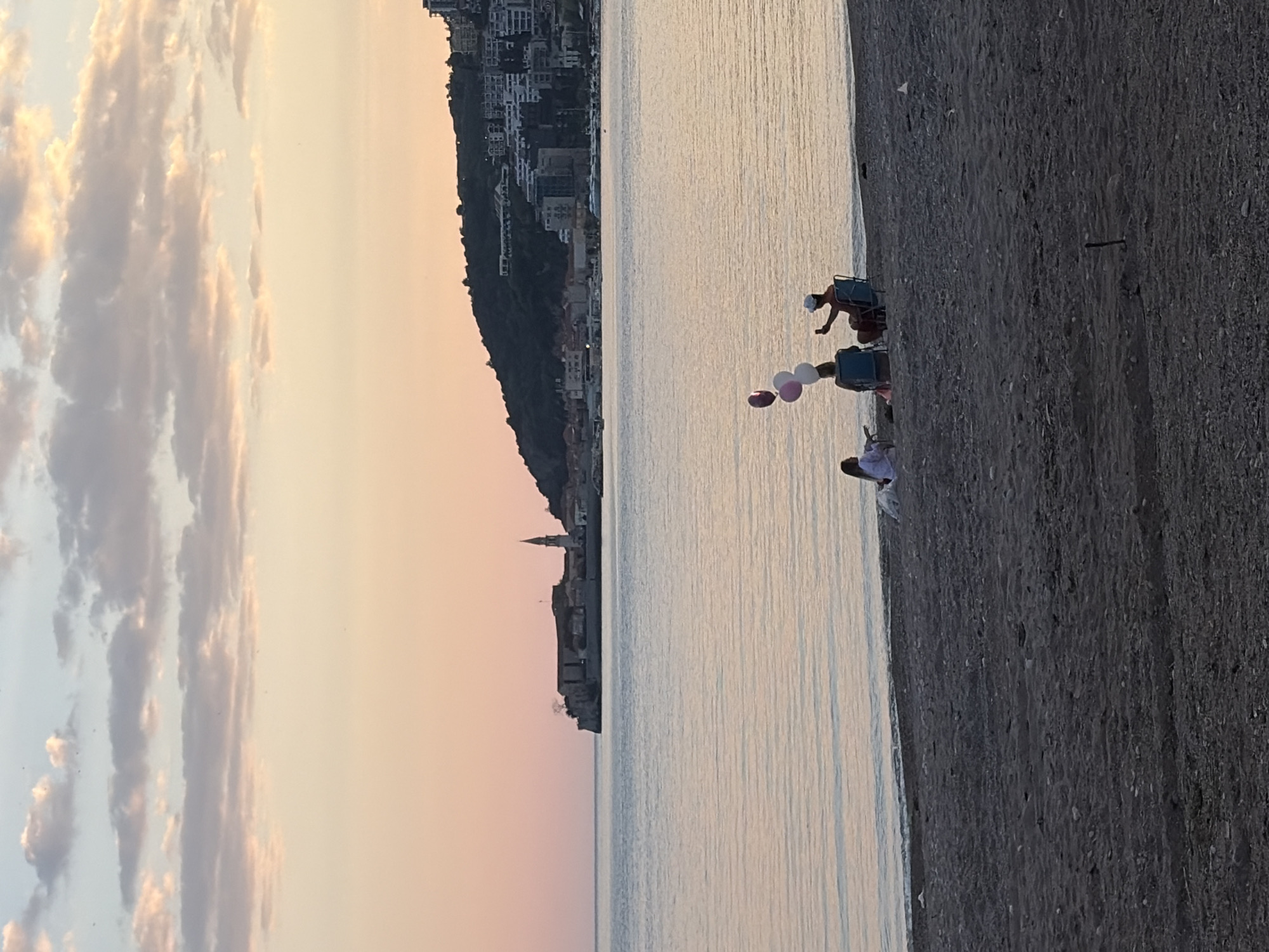 The photograph shows a coastal scene in Budva, Montenegro, at sunset. The sky is streaked with pink and pale orange light, partly obscured by clouds. To the left of the frame, a hillside rises, topped with buildings, and a church spire is visible. The calm sea reflects the colors of the sky. On the right, a dark, pebbly beach runs along the water's edge. Several people are seated in chairs on the beach, facing the water. One person holds balloons.
