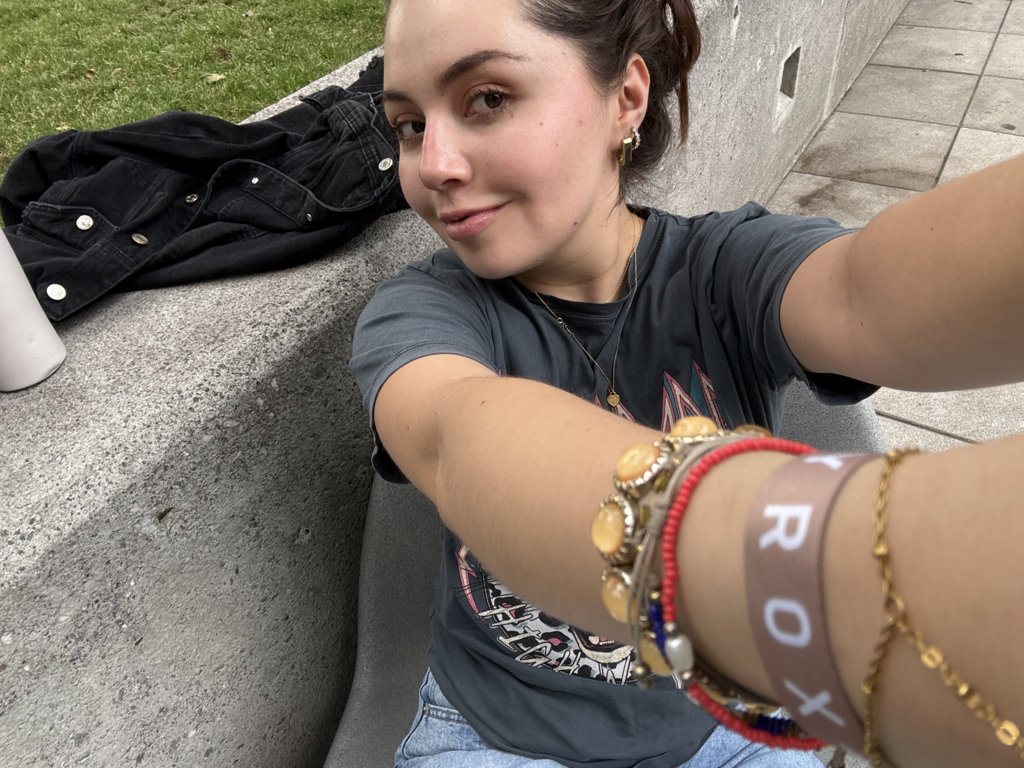 A selfie taken by a young woman with light skin and brown hair in a ponytail, sitting on a concrete bench. She wears a gray t-shirt, blue jeans, and several bracelets on her right wrist. A black denim jacket and a white container rest on the bench beside her. The background includes a concrete wall and paved sidewalk, suggesting an urban location, possibly in Mexico City, Mexico.