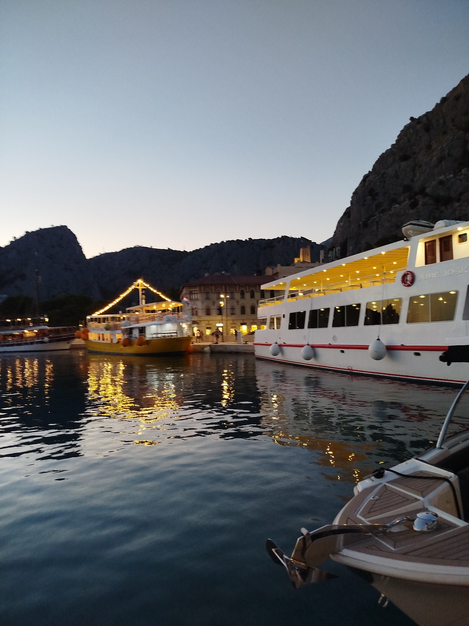 This is a vertical, eye-level, outdoor photograph taken at dusk in the city of Omiš, Croatia. The scene depicts a harbor with several boats docked. In the foreground, the bow of a smaller white boat with a polished metal anchor is visible. To the left and center of the frame are two larger boats. One is yellow, with its lights illuminated, and features a string of lights draped across its top deck, casting a reflection on the water. The other boat is white with red trim and numerous windows. The water in the harbor is calm, reflecting the lights from the boats and the sky. In the background, buildings with illuminated windows line the waterfront, and steep, rocky hills rise behind them. The sky is a gradient of light blue at the top to a pale orange near the horizon, indicating twilight.
