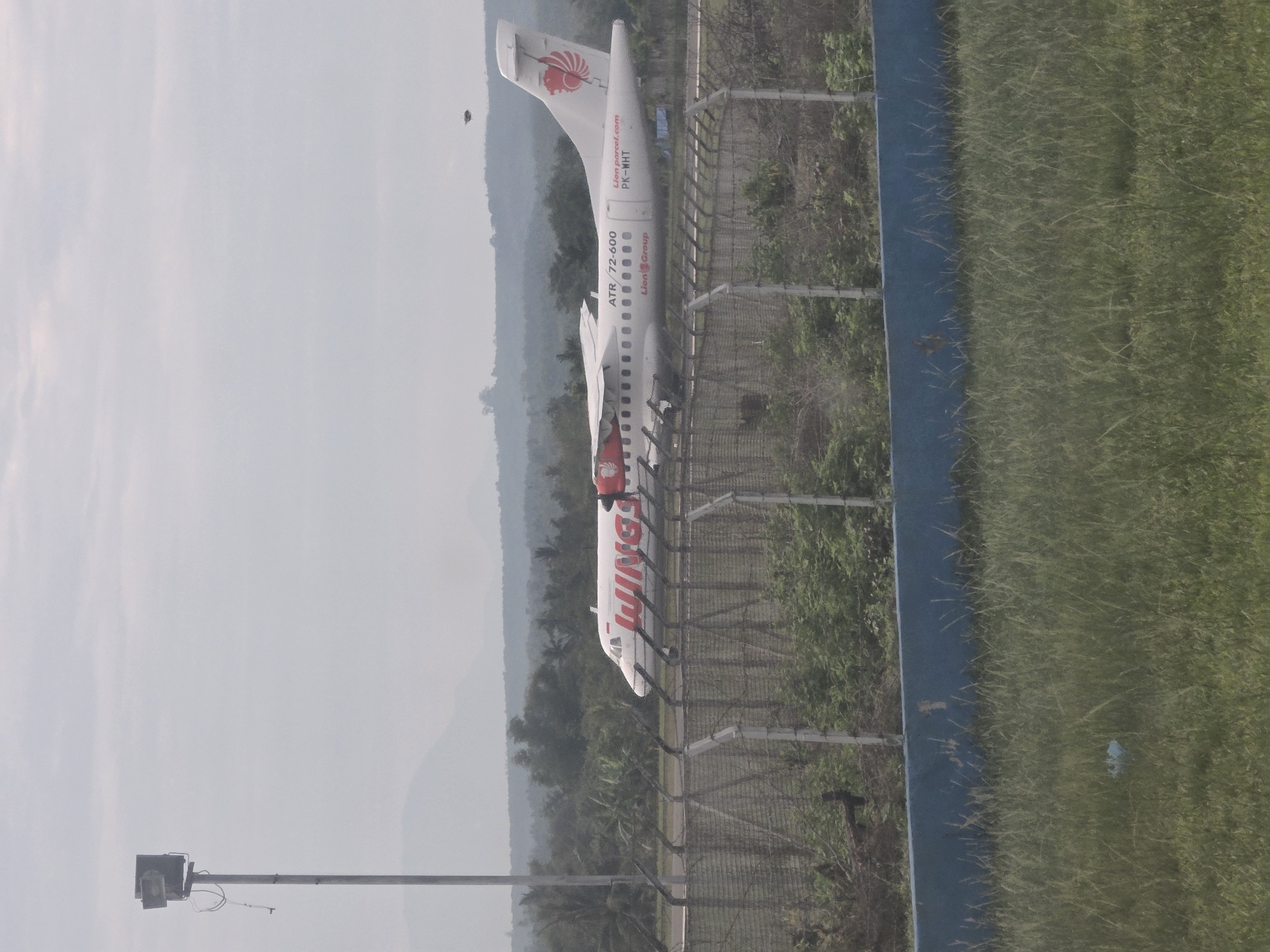 An ATR 72-600 aircraft, registered as PK-WHT and operated by Wings Air (a subsidiary of Lion Group), is parked at Pintomakmur Airport in Indonesia. The aircraft is painted white with red "WINGS" lettering on the fuselage and a red lion's head logo on the tail. The background includes mountains and a fence, separating the runway from grassy areas. A tall pole with a device is seen in the bottom left corner.