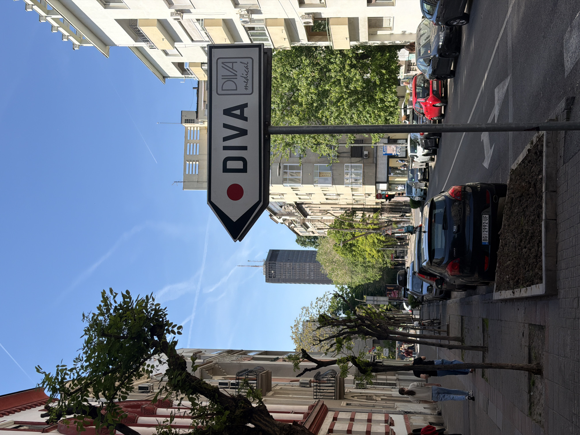 The image is a street view in Belgrade, Serbia. A sign points towards "DIVA medical" with a red dot on the left. Parked cars line the right side of the street. Two people walk on the sidewalk in the same direction as the sign points. Trees are planted alongside the sidewalk. Buildings line both sides of the street. In the distance is the Belgrade Tower. The sky is blue with contrails from airplanes.