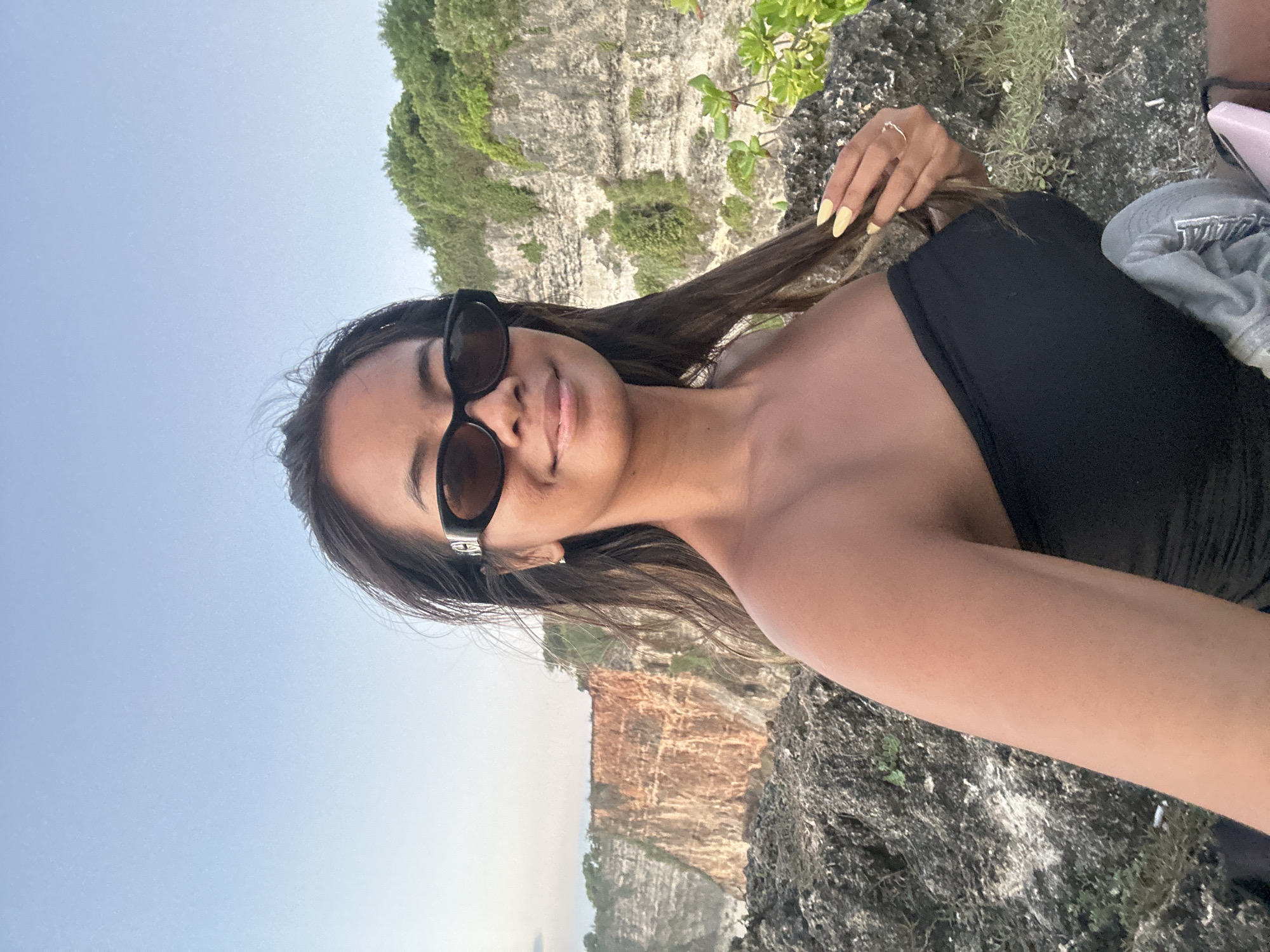 In the city of Kuta Selatan, Indonesia, a young woman of East Asian descent with long, dark hair, wearing black sunglasses and a black strapless top, poses for a selfie. She is positioned on a rocky outcrop overlooking a cliff face. The cliff is multi-layered, with distinct horizontal strata, and appears to be made of reddish-brown sedimentary rock. Lush green vegetation covers the top of the cliff and some parts of its face. The sky above is a clear, pale blue. The foreground shows the woman's arm and hand, which has bright yellow nail polish and a delicate ring on one finger. She is touching her hair with her right hand. Behind her, to the right, is a small amount of green foliage growing from the rocks. A grey cap is visible on her lap, and a pink object, possibly a phone case, rests beside it. The overall scene suggests an outdoor setting, likely during the daytime, with the natural landscape of the Indonesian coast as the backdrop.