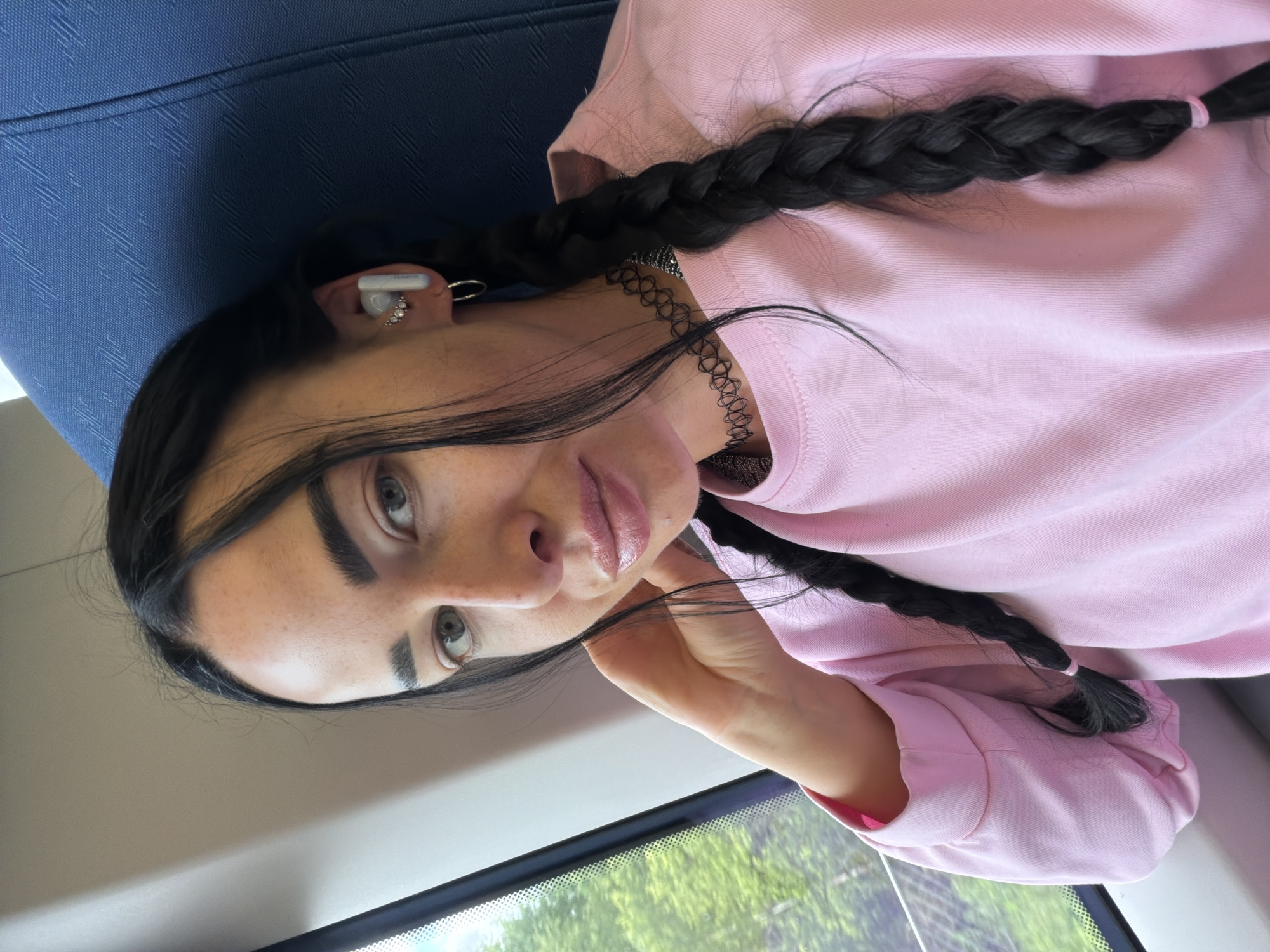 This is a portrait of a young woman indoors, possibly on a train, given the blue patterned seat and window view. She has long, dark hair styled in two braids, and is wearing a pink sweatshirt and a black choker necklace. She also has white earbuds in her ears and several rings on her fingers, including a small diamond stud in her left ear. Her expression is neutral, and she is looking towards the camera. The lighting suggests it is daytime, and the blurry greenery outside the window indicates a possible outdoor landscape. No visible text is present in the image. The location context provided is Ушаки, Russia.