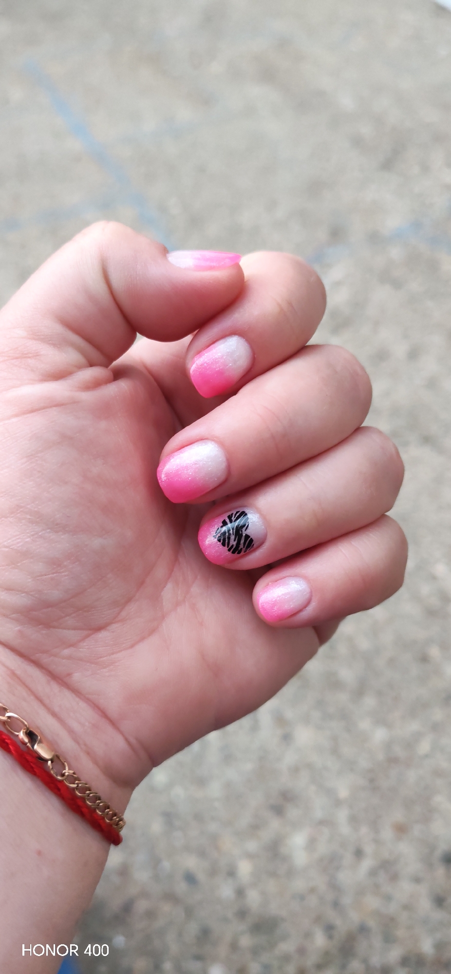 The image shows a close-up of a hand with a manicure. The nails are short and appear to be acrylic or gel. They have a gradient of pink and white, with a black zebra print design on one of the nails. A red string bracelet with a gold clasp is visible on the wrist. The background is blurry and appears to be an outdoor setting with concrete or pavement. The bottom left corner of the image displays "HONOR 400", which is likely a model identifier for the camera used to take the photo. There is no discernible location in the city of Noviy, Russia.